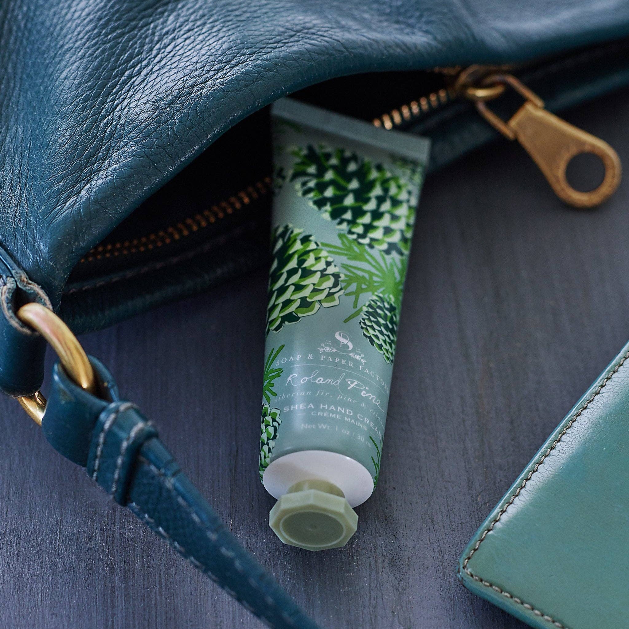 A tube of Roland Pine Shea Butter Hand Cream is partially pulled out from a blue leather purse, with a green and white patterned design on the tube.