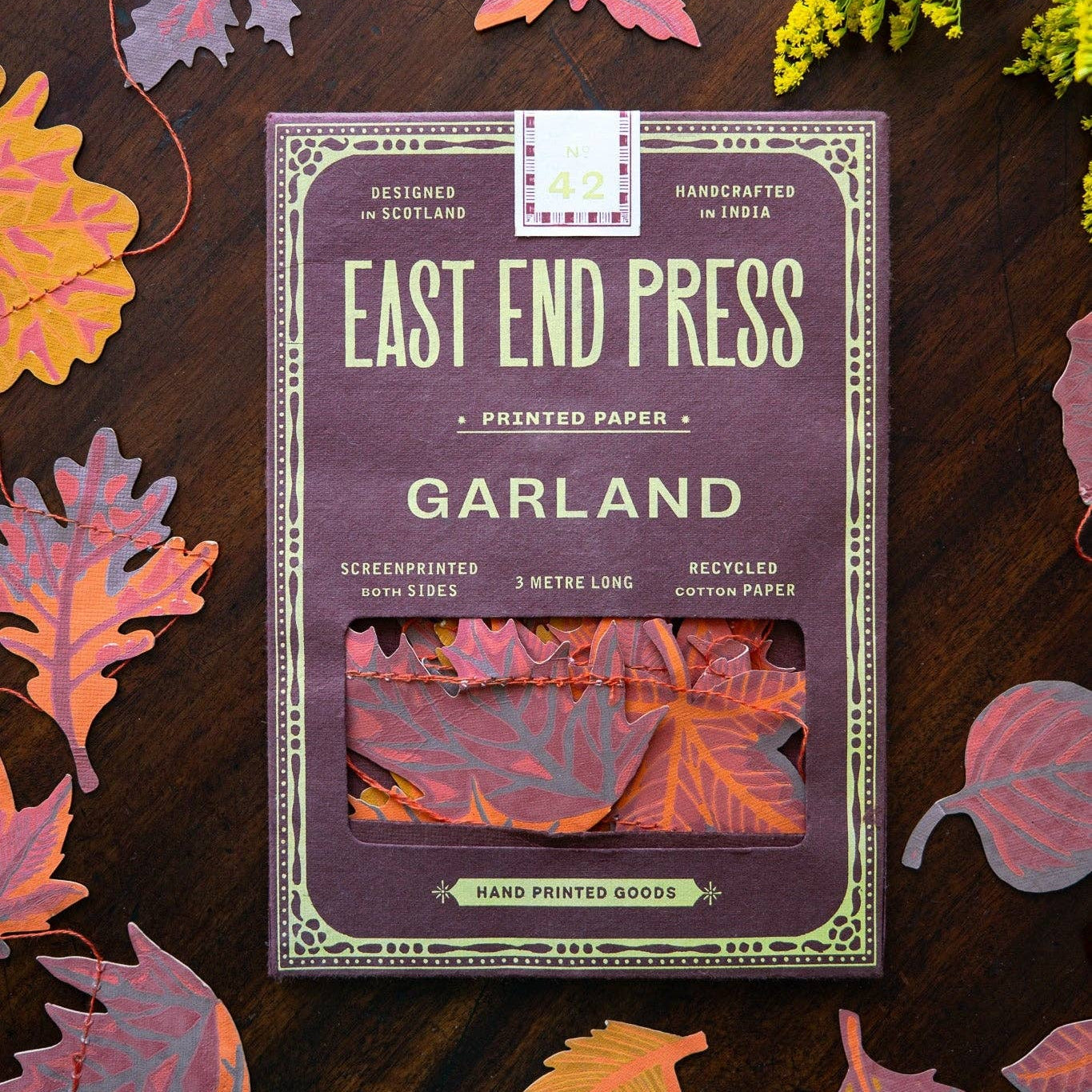 A package of East End Press Garland printed paper sheets lies on a dark wooden surface surrounded by colorful autumn leaves and small yellow flowers. The package is mauve-purple with cream text and border details, and a window near the bottom shows folded paper samples in pink, orange, and red tones.
