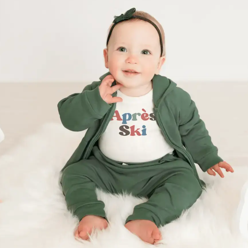 Baby wearing a green outfit with a colorful 'Après Ski' shirt on a white background