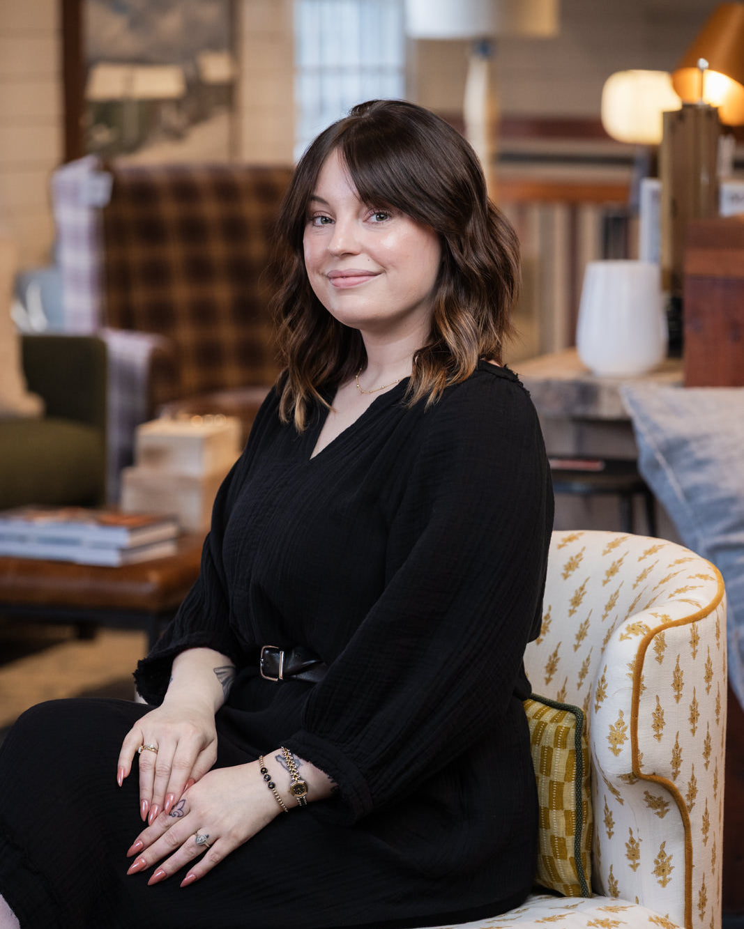 A woman with shoulder-length brown hair sits in a patterned armchair wearing a black long-sleeve dress with a thin belt. The setting is a warm, softly lit interior with home decor in the background.