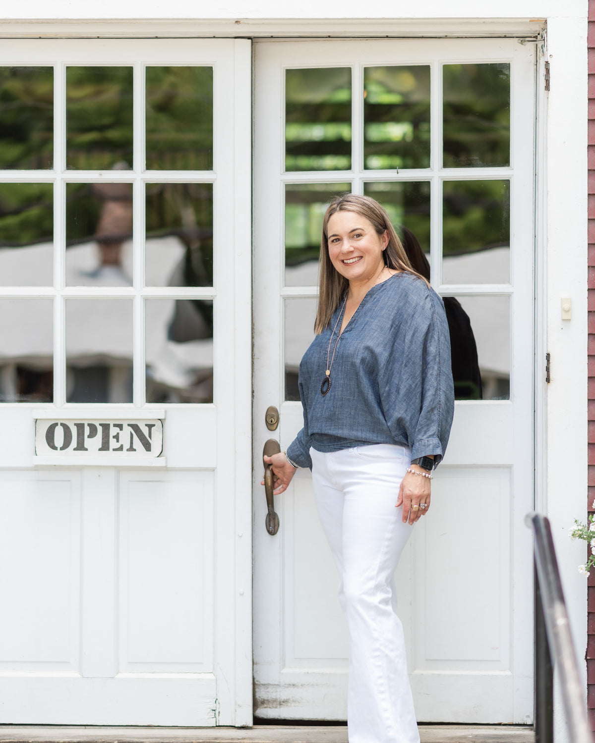 A woman stands in front of a white storefront door with glass panes, wearing a blue long-sleeve blouse and white pants. The door has an 'OPEN' sign and a metal handle.
