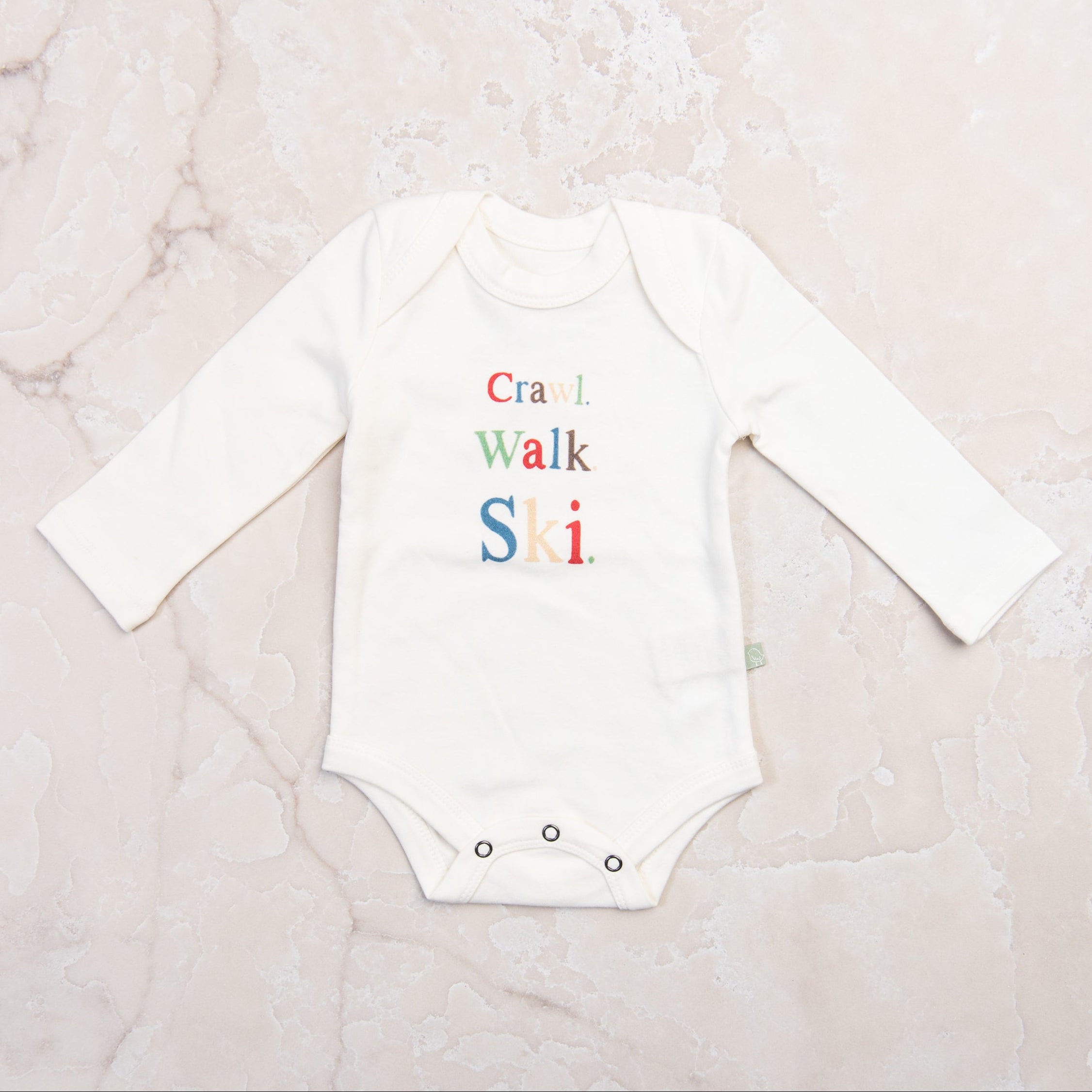 Baby Long Sleeve Graphic Organic Bodysuit in Crawl Walk Ski on a marble table at Addison West