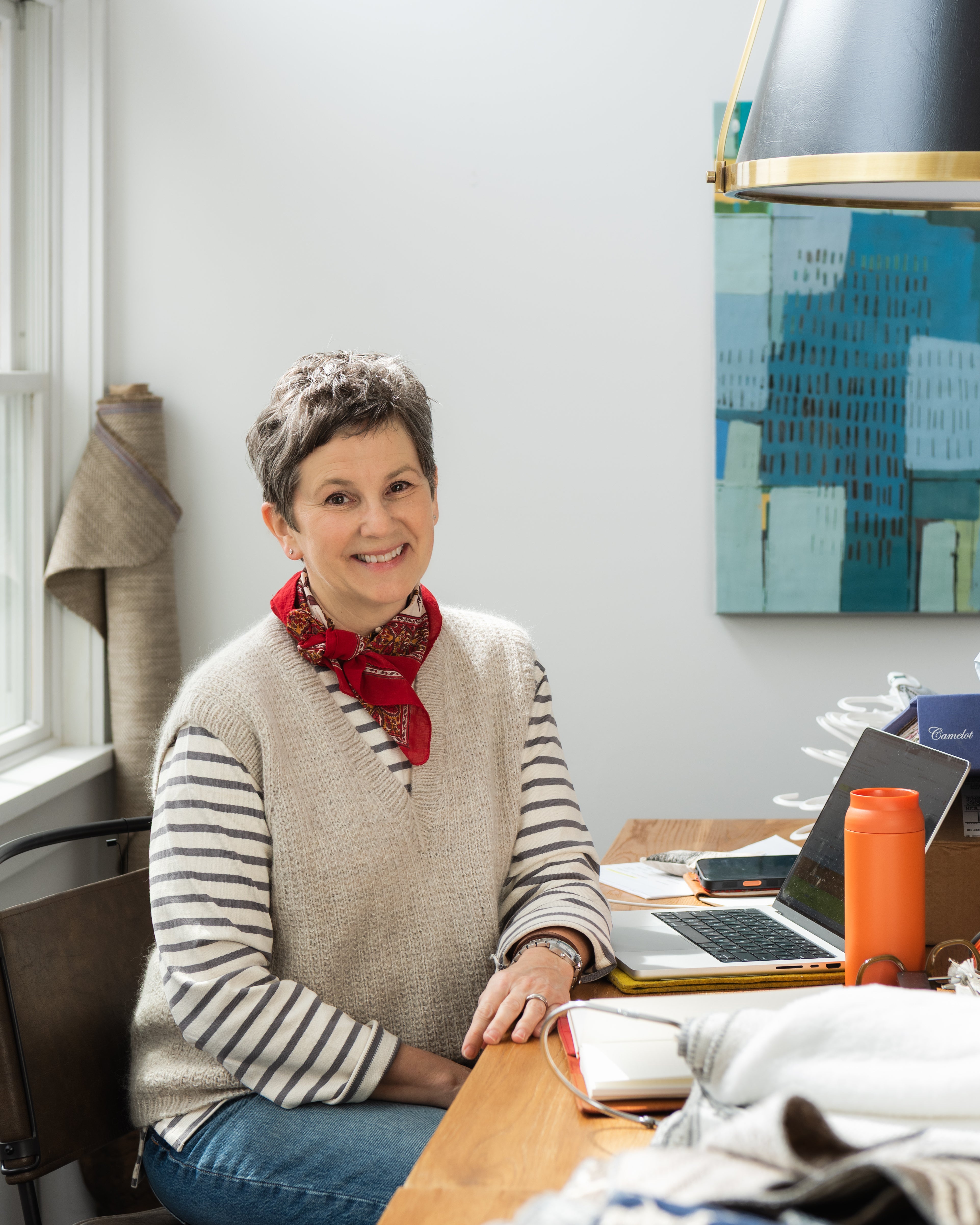 A smiling woman with short gray hair sits at a wooden desk in a bright room, wearing a beige knit sweater vest over a black-and-white striped long-sleeve shirt, blue jeans, and a red patterned neck scarf. A laptop, orange bottle, and patterned fabric are visible on the desk.