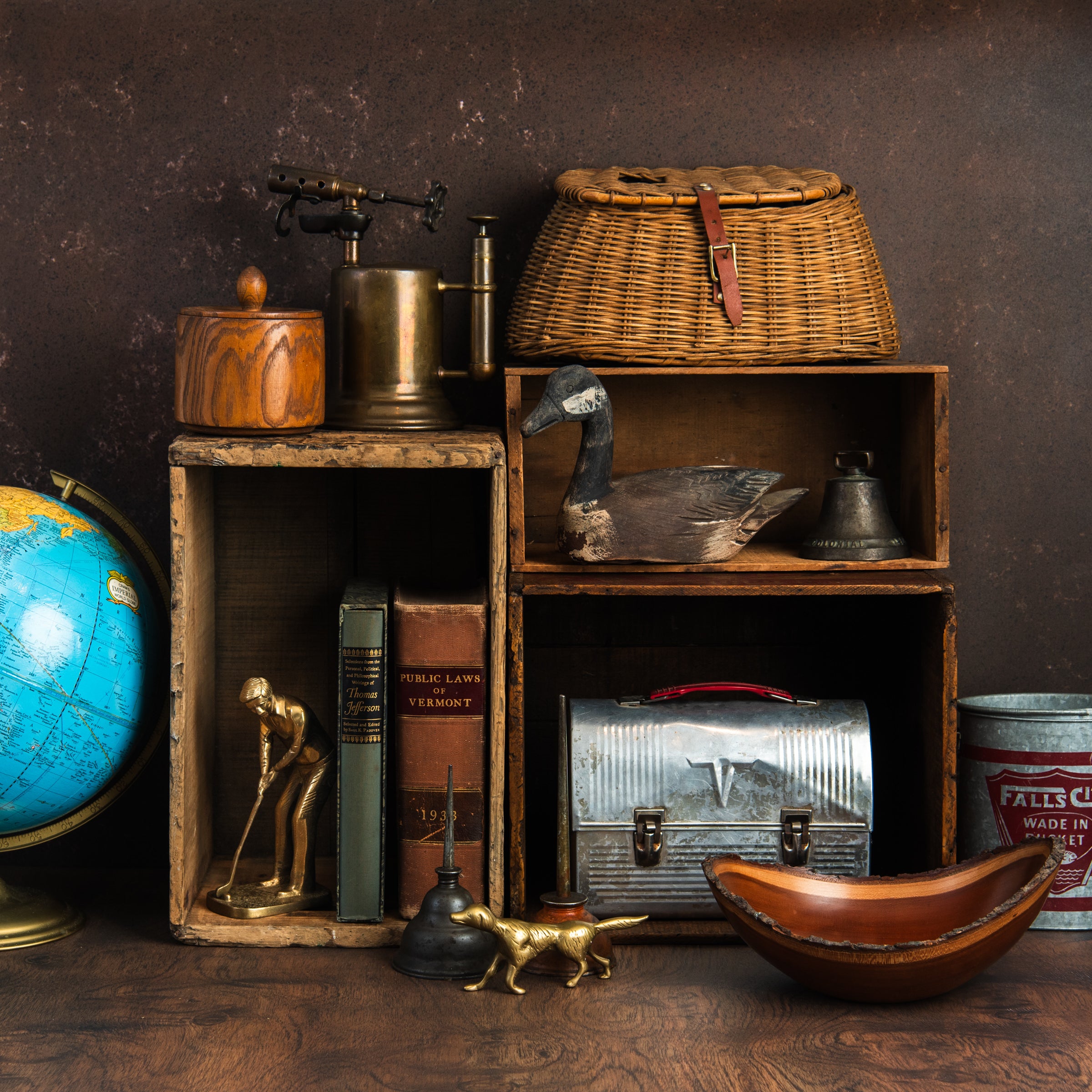Vintage collection including fishing creel, brass blow torch, wood humidor, globe, decoy, brass golfer, brass dog, vintage oil cans, lunch box, and books on a wood table at Addison West