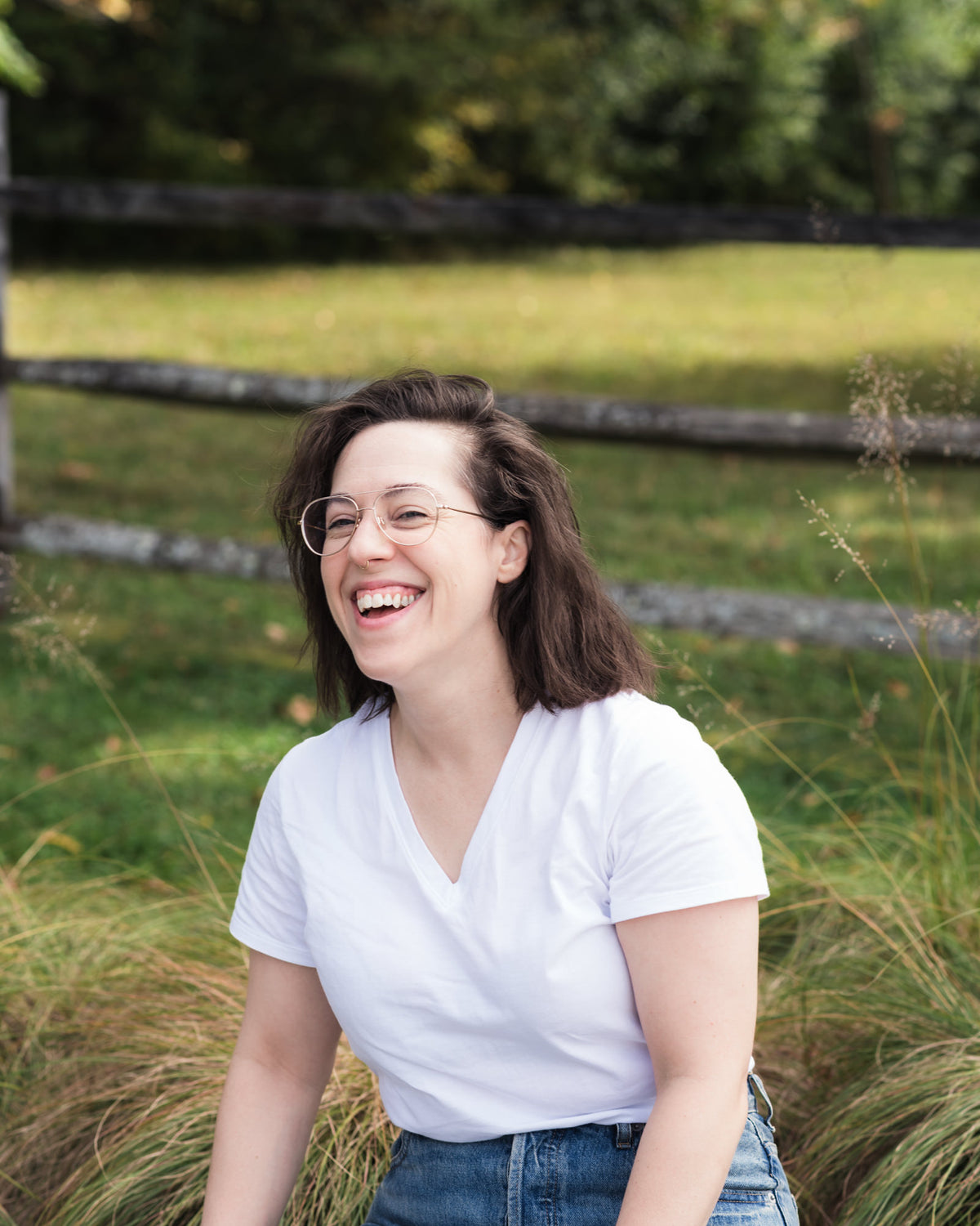 A woman with brown hair and glasses, wearing a white V-neck t-shirt and blue jeans, sitting outdoors in a grassy field with a wooden fence in the background.