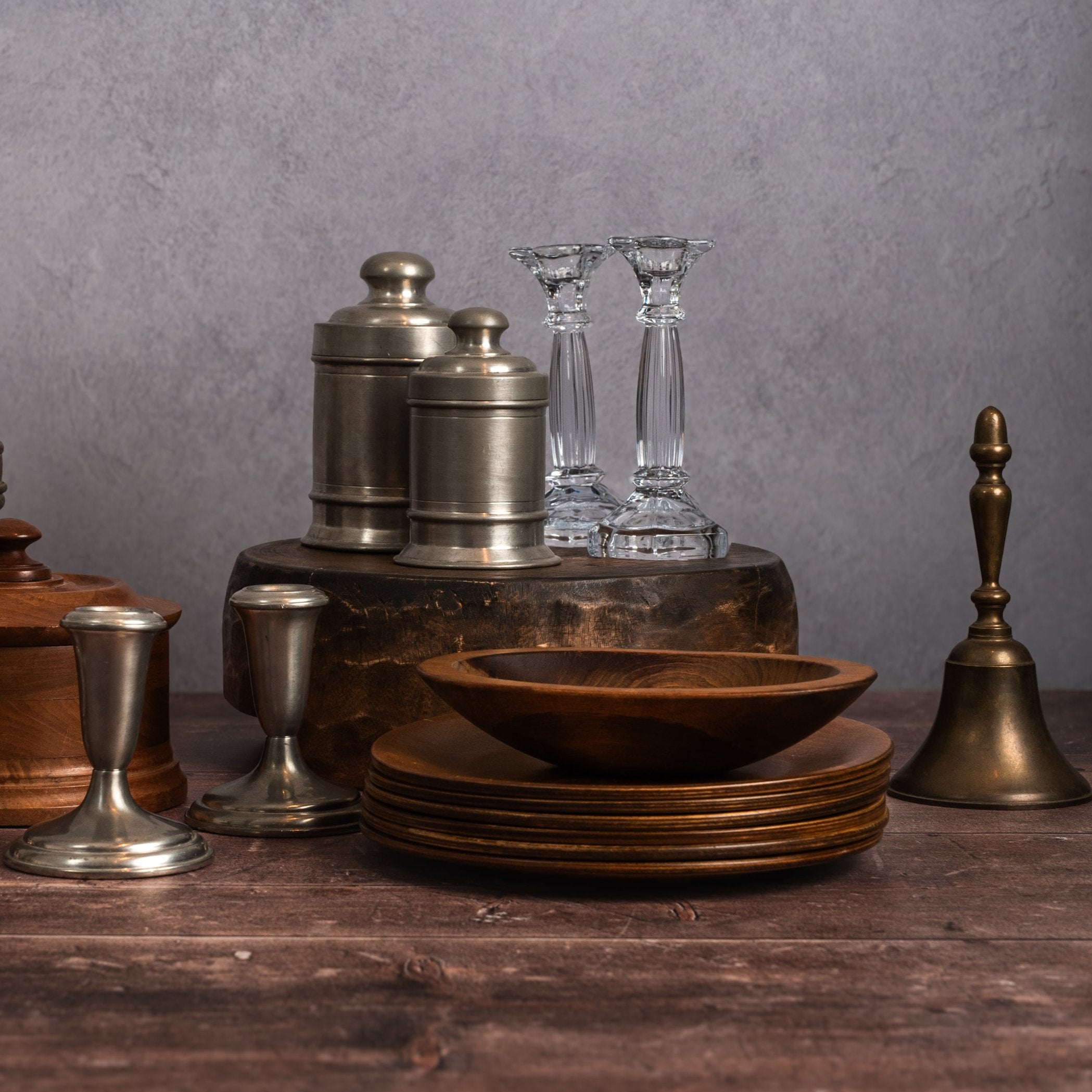 Grouping of vintage glass candlesticks, brass bell, pewter candlesticks, silver jars, and wood bowl and plates on a wood table and white background at Addison West.