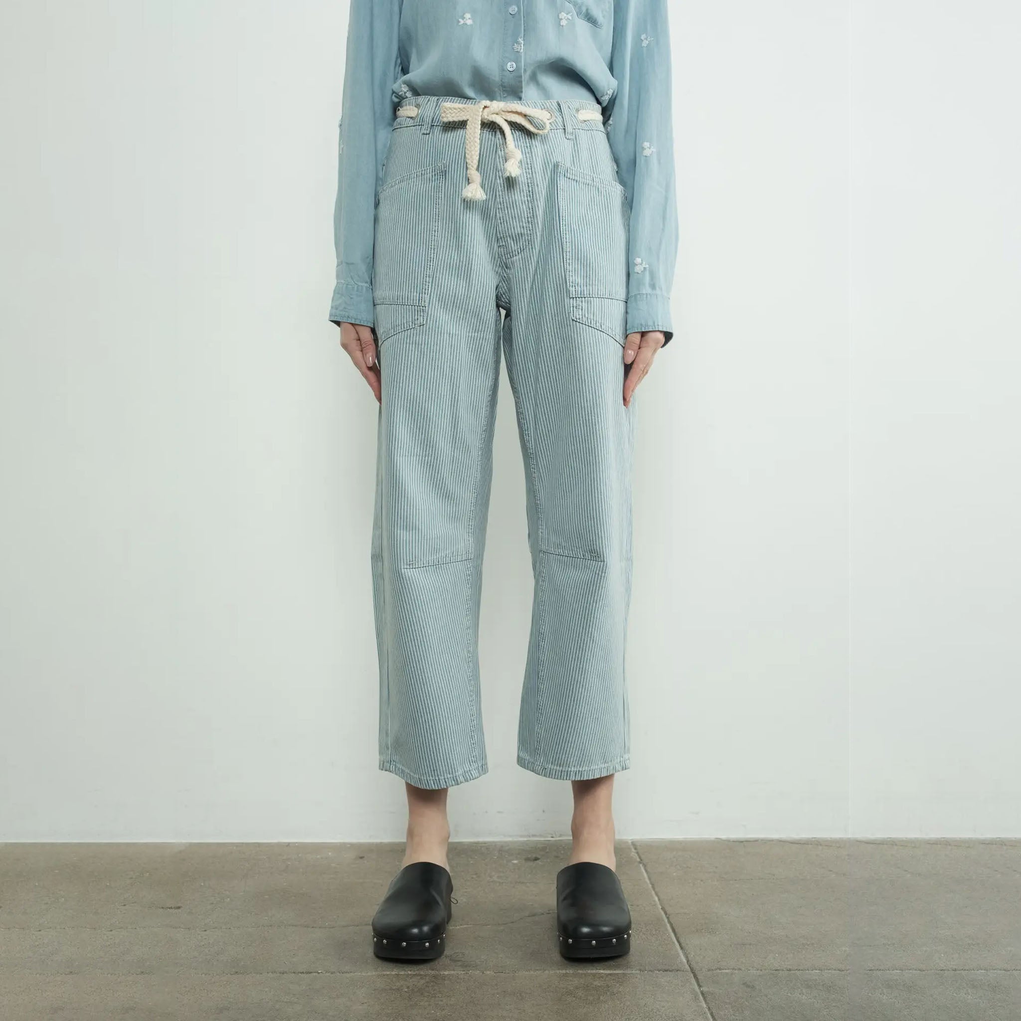 Woman wearing a light blue shirt and Aaron and Amber striped denim barrel pants standing against a white wall.
