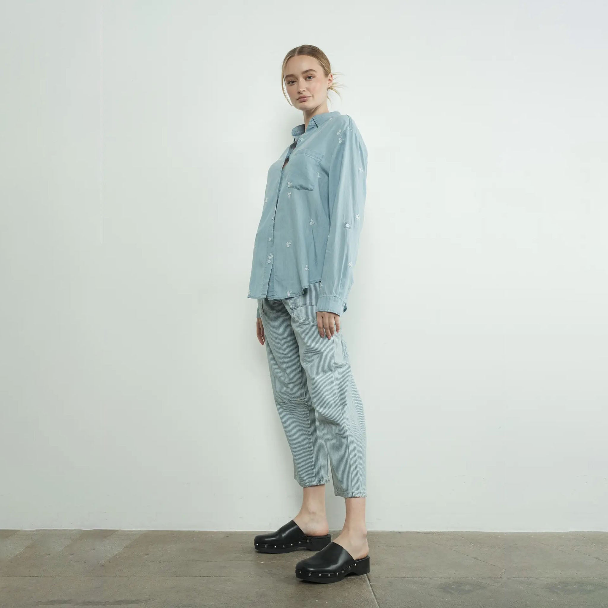 Woman wearing a light blue shirt and Aaron and Amber striped denim barrel pants standing against a white wall.