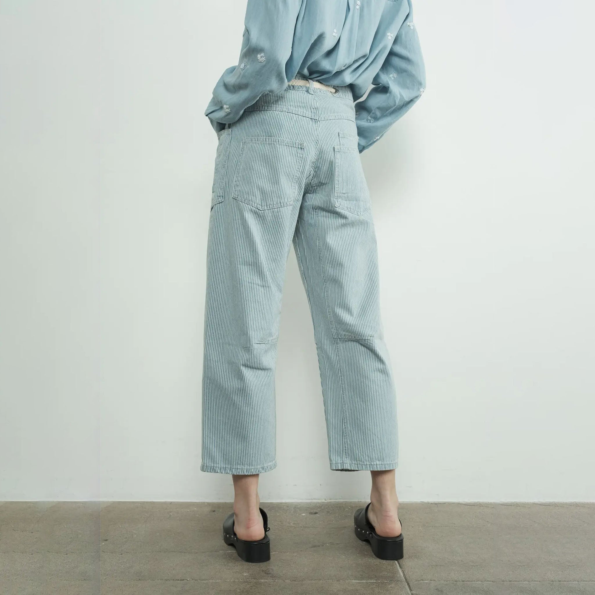 Woman wearing a light blue shirt and Aaron and Amber striped denim barrel pants standing against a white wall.