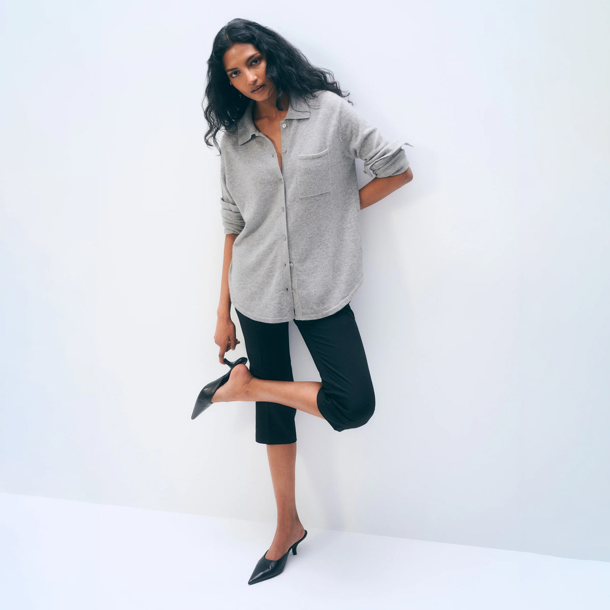 A woman posing in a light gray button-down shirt, black cropped capri pants, and black pointed-toe mule heels against a clean white studio background.