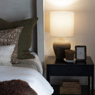 Upholstered bed with pillows next to black bedside table and clay pot lamp with white shade
