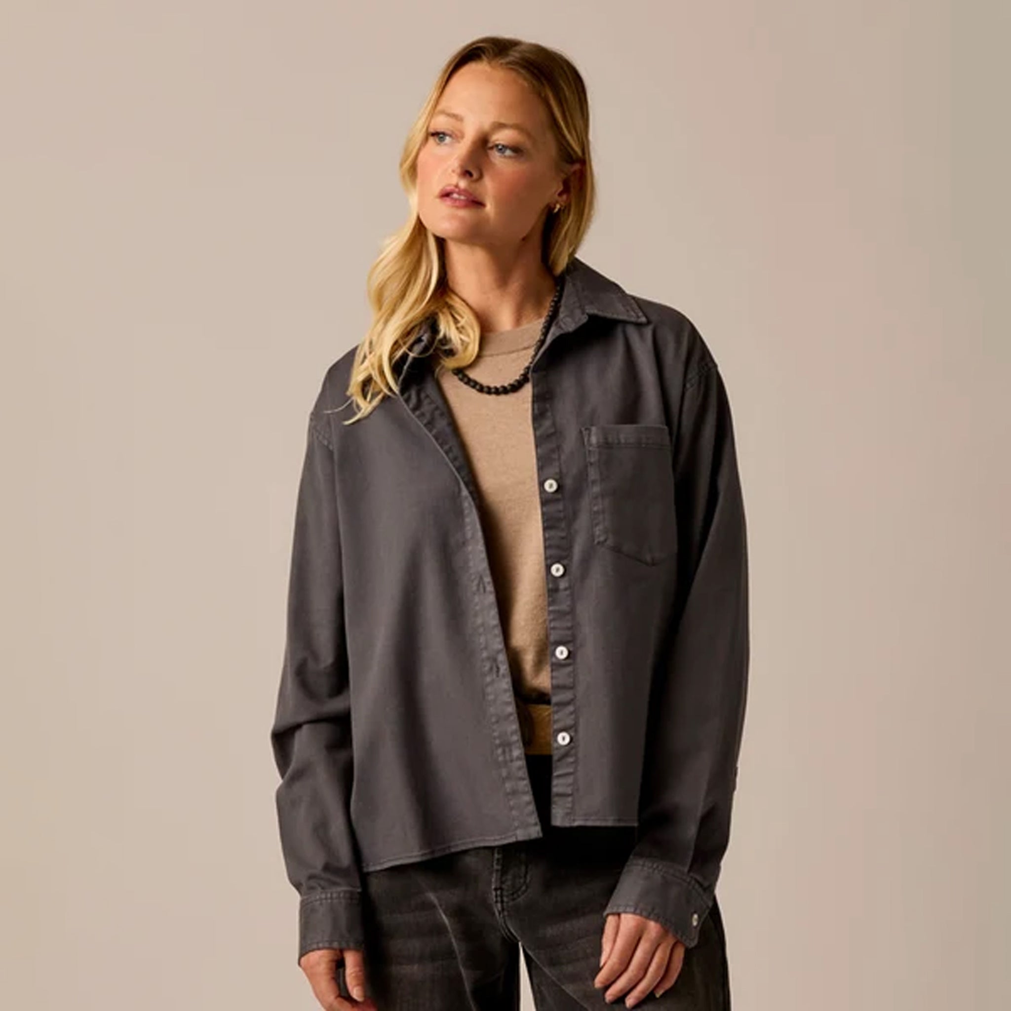 A woman modeling a dark charcoal gray button-up shirt with a chest pocket, worn open over a beige knit top and dark jeans, set against a solid light beige studio background.