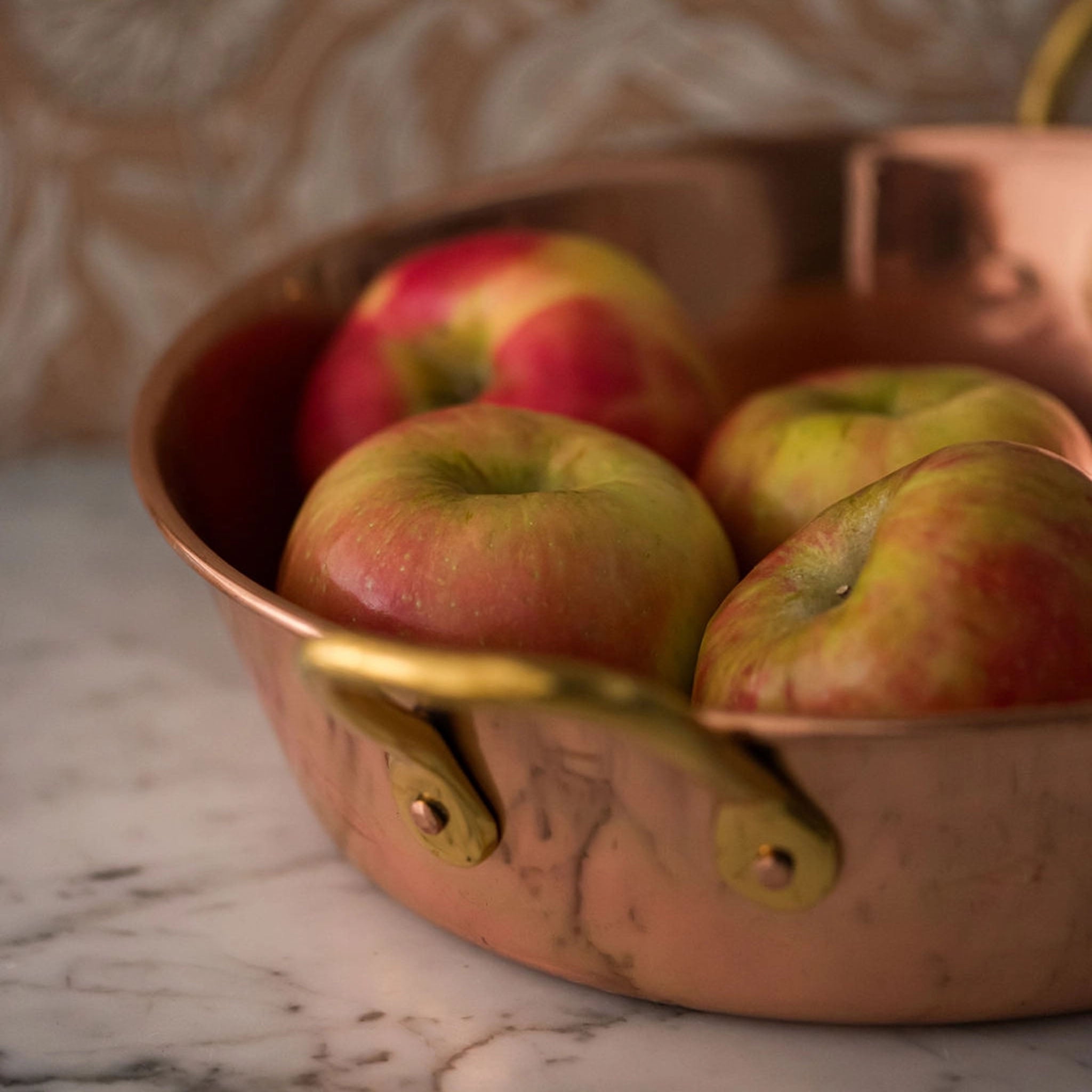 Copper pan with brass handle with apples inside on a marble counter
