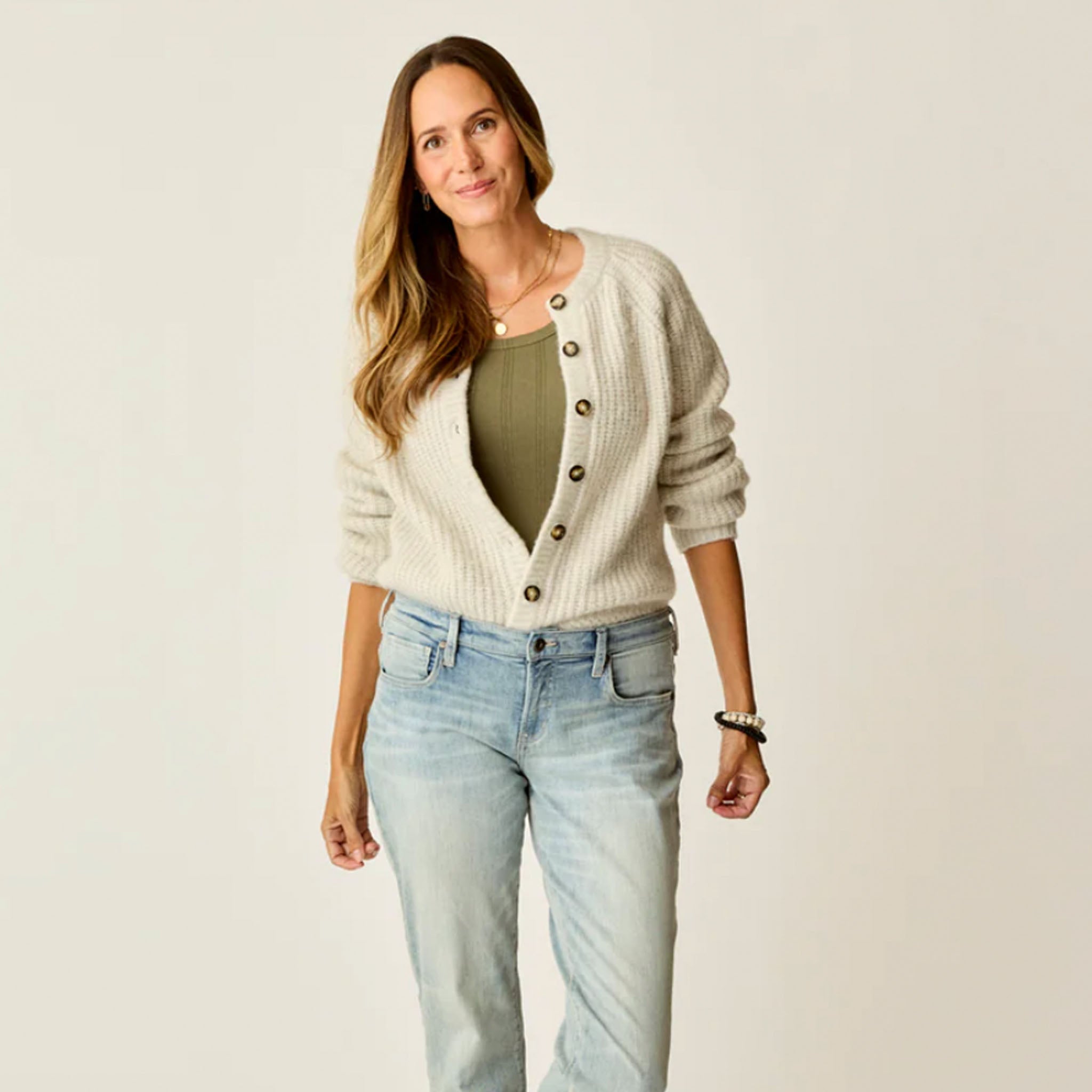 Woman wearing a cream cardigan, green top, and light blue jeans on a plain background