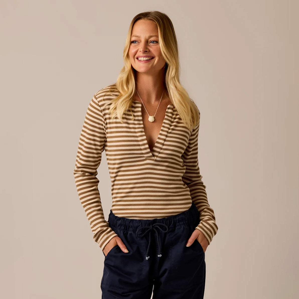 A smiling blonde woman wearing a brown and beige striped long-sleeve wrap top and navy drawstring pants, standing against a solid beige studio background.