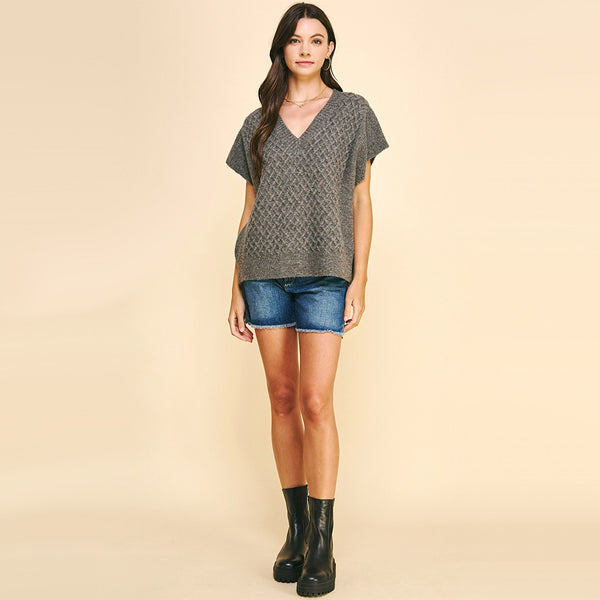 Brunette model wearing Pinch Crochet Sweater Vest in Charcoal with boots and jean shorts in front of a yellow background