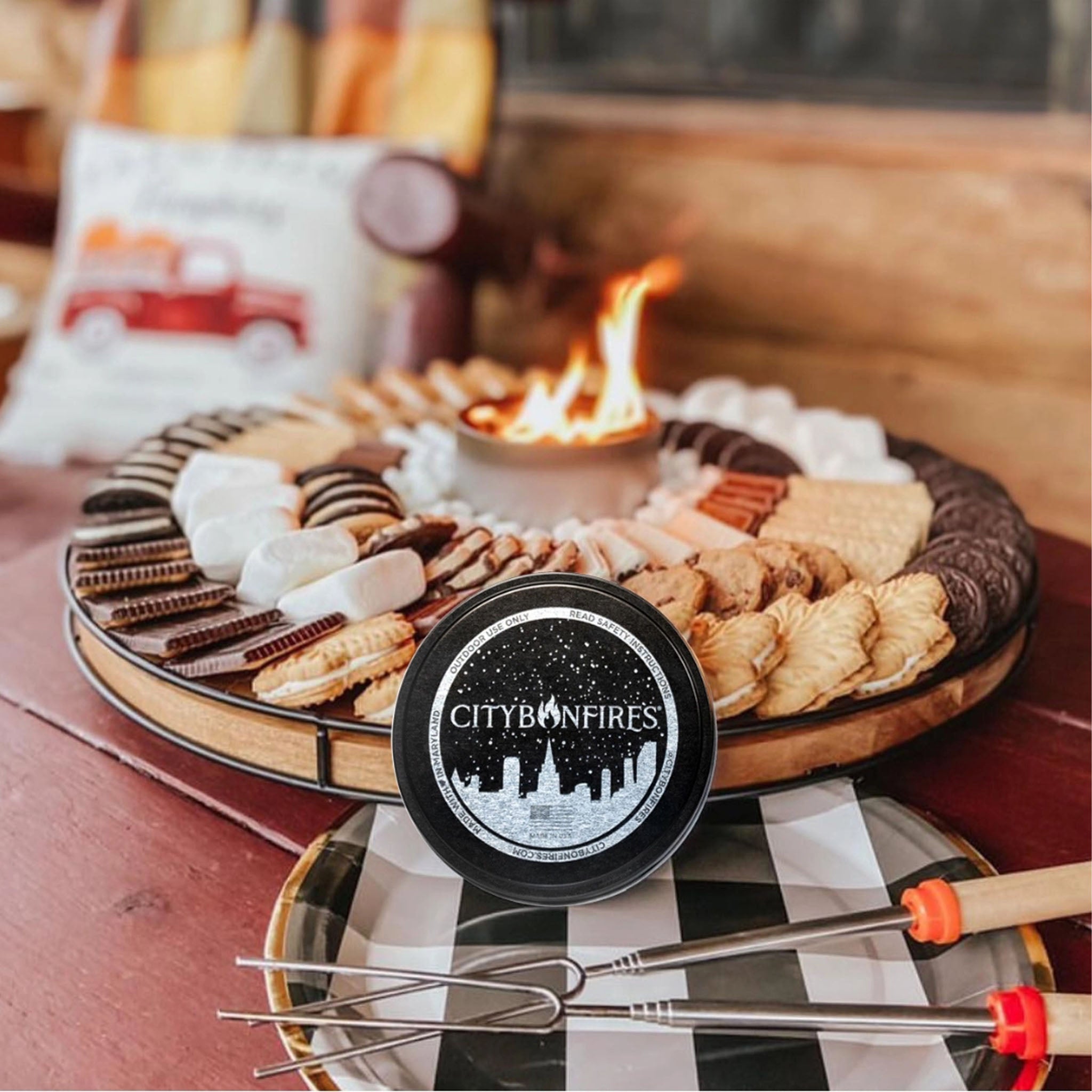 Fire pit with marshmallows, cookies, and a City Bonfires branded container on a checkered tablecloth.