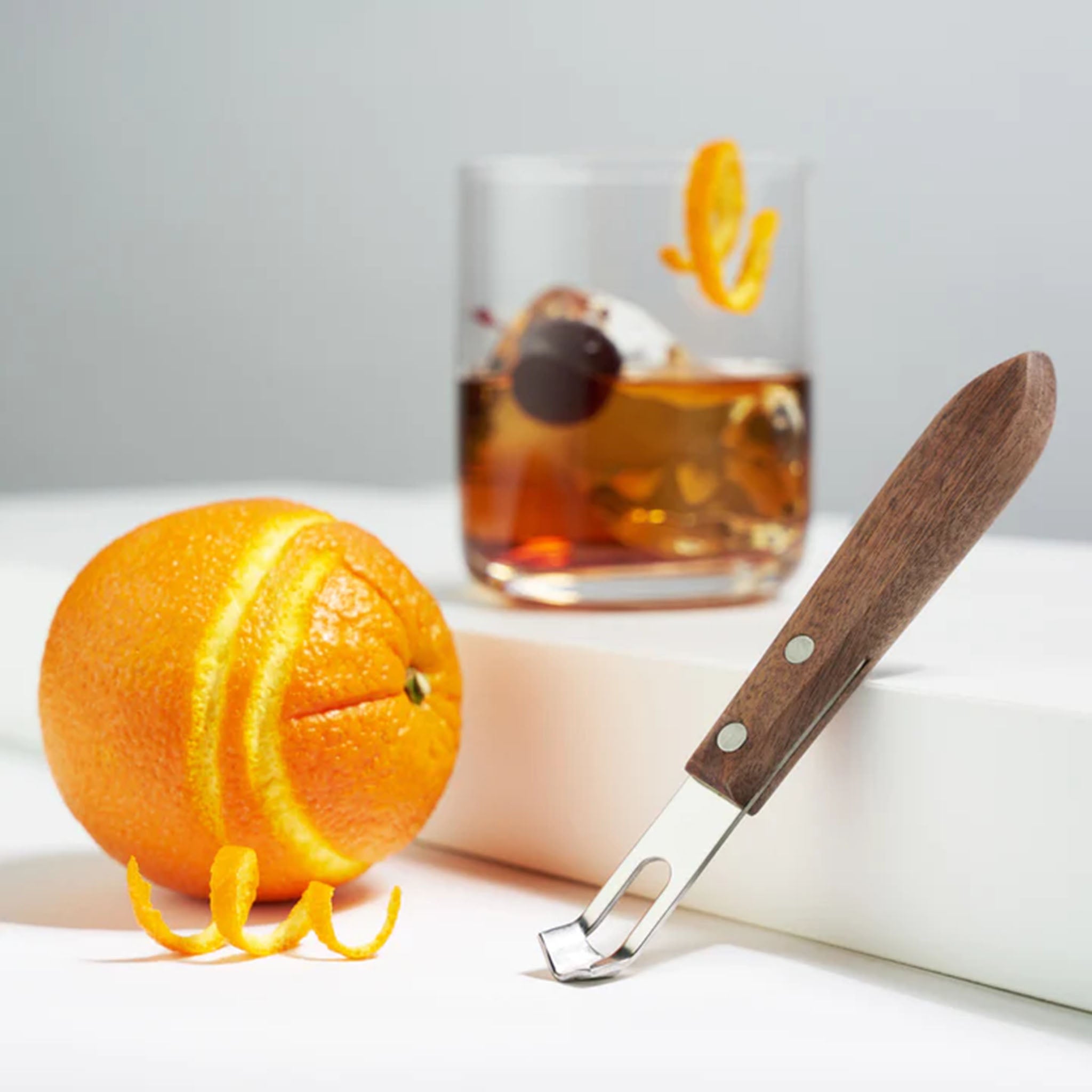 Glass with ice and liquor on a white counter next to an orange and Stainless steel Channel Knife with Walnut Handle