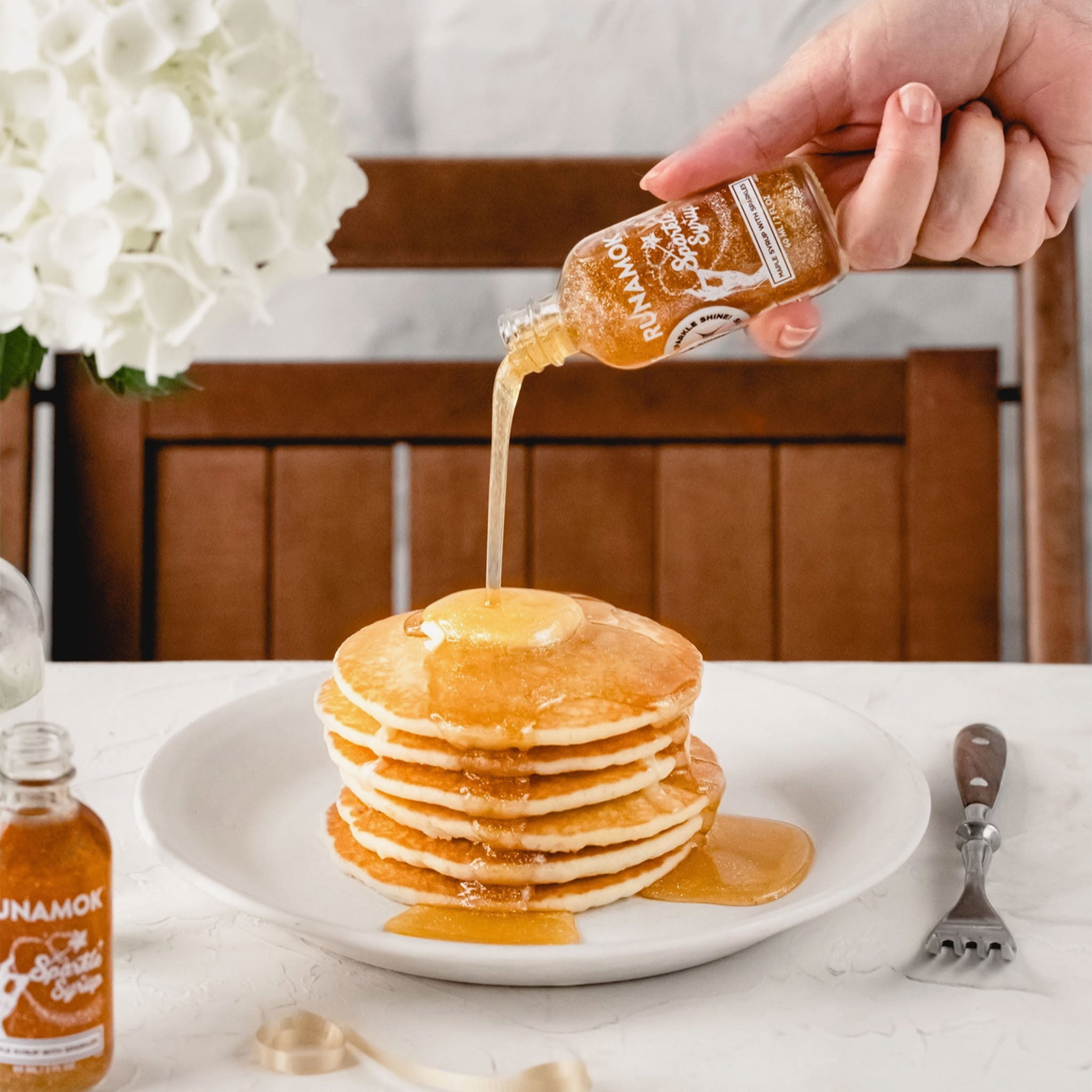 Hand pouring Runamok sparkle maple syrup over pancakes on a table with a white table cloth