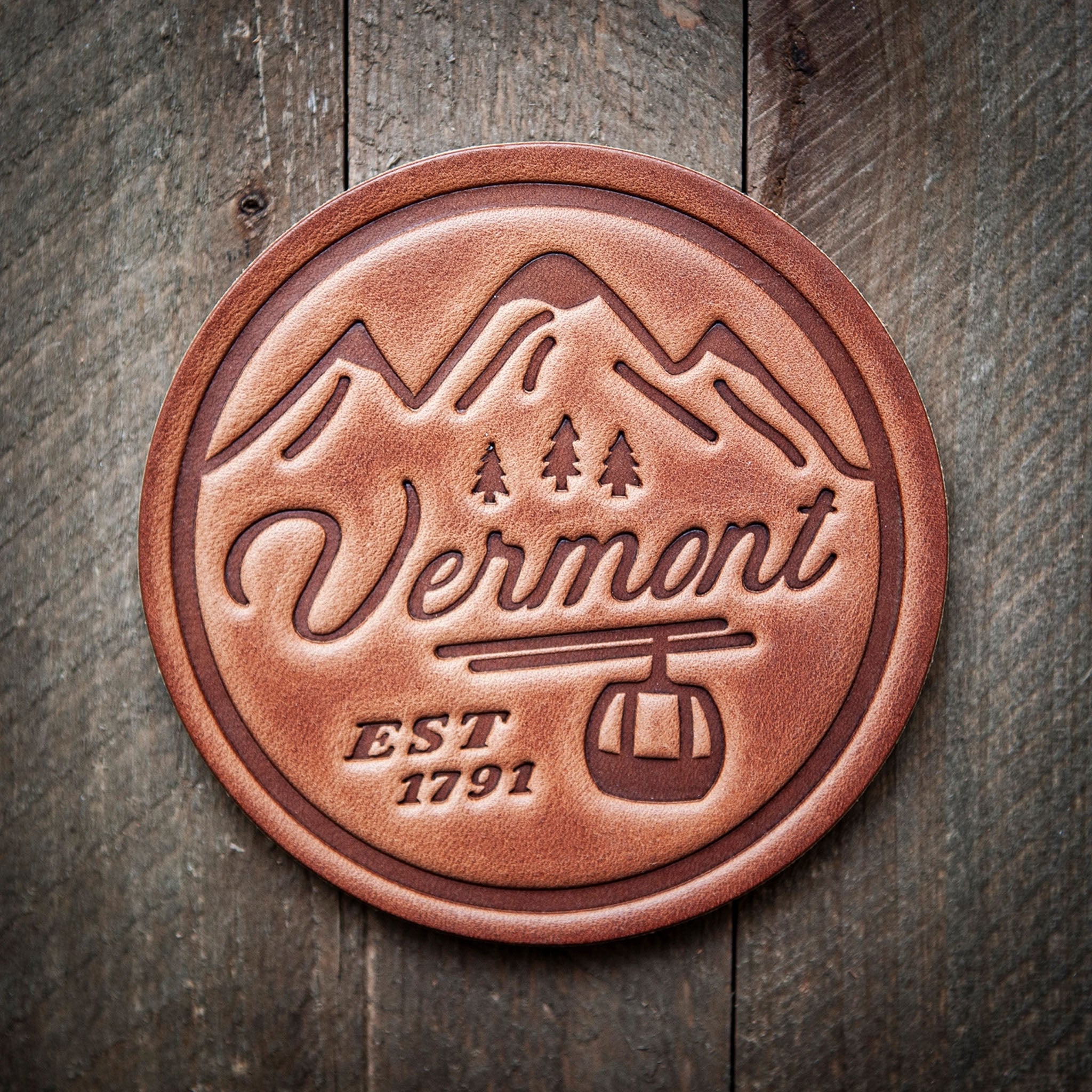 A brown leather coaster with embossed text and a ski gondola design, on a wooden background.