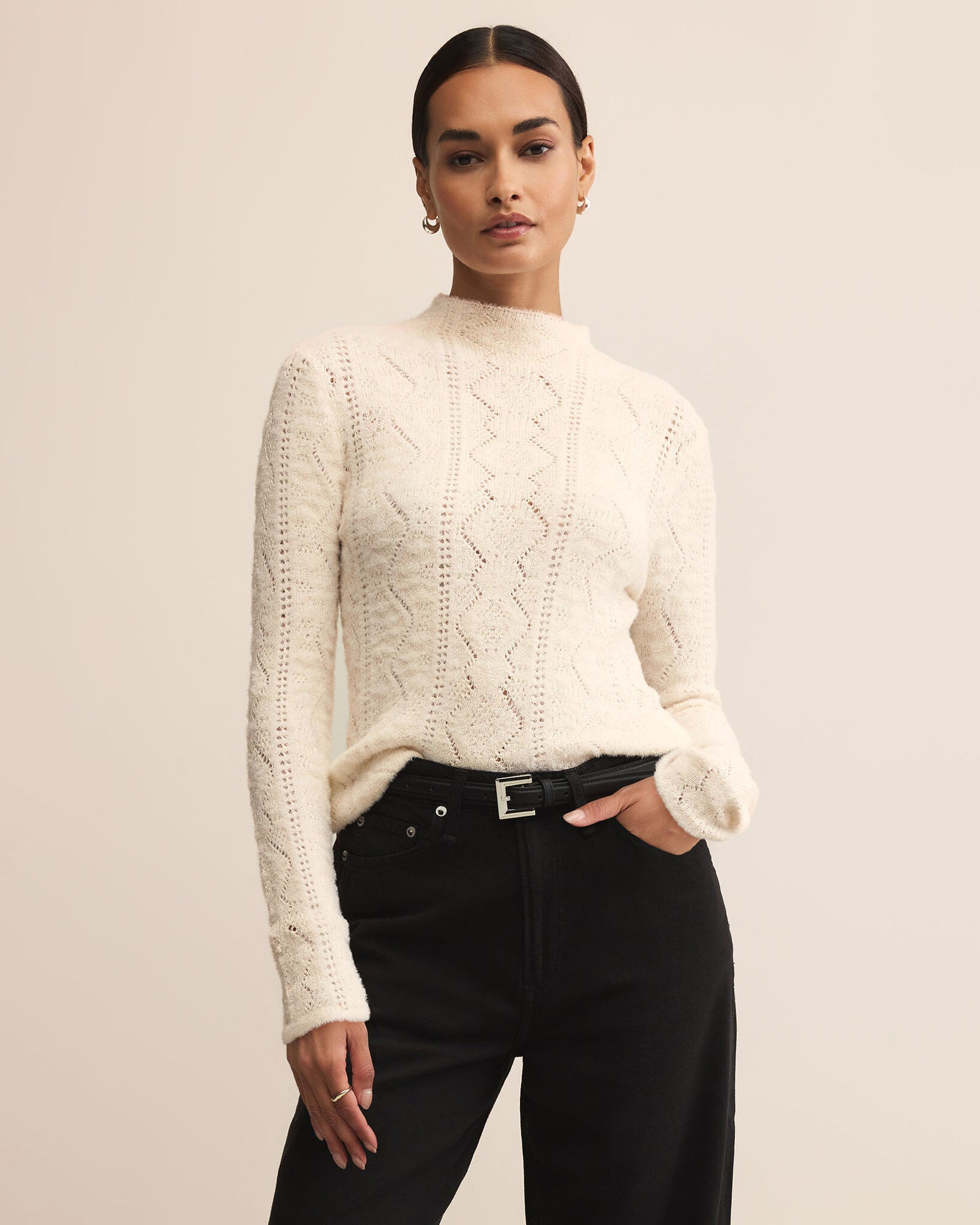 Woman wearing cream colored sweater with dark pants on a plain background.