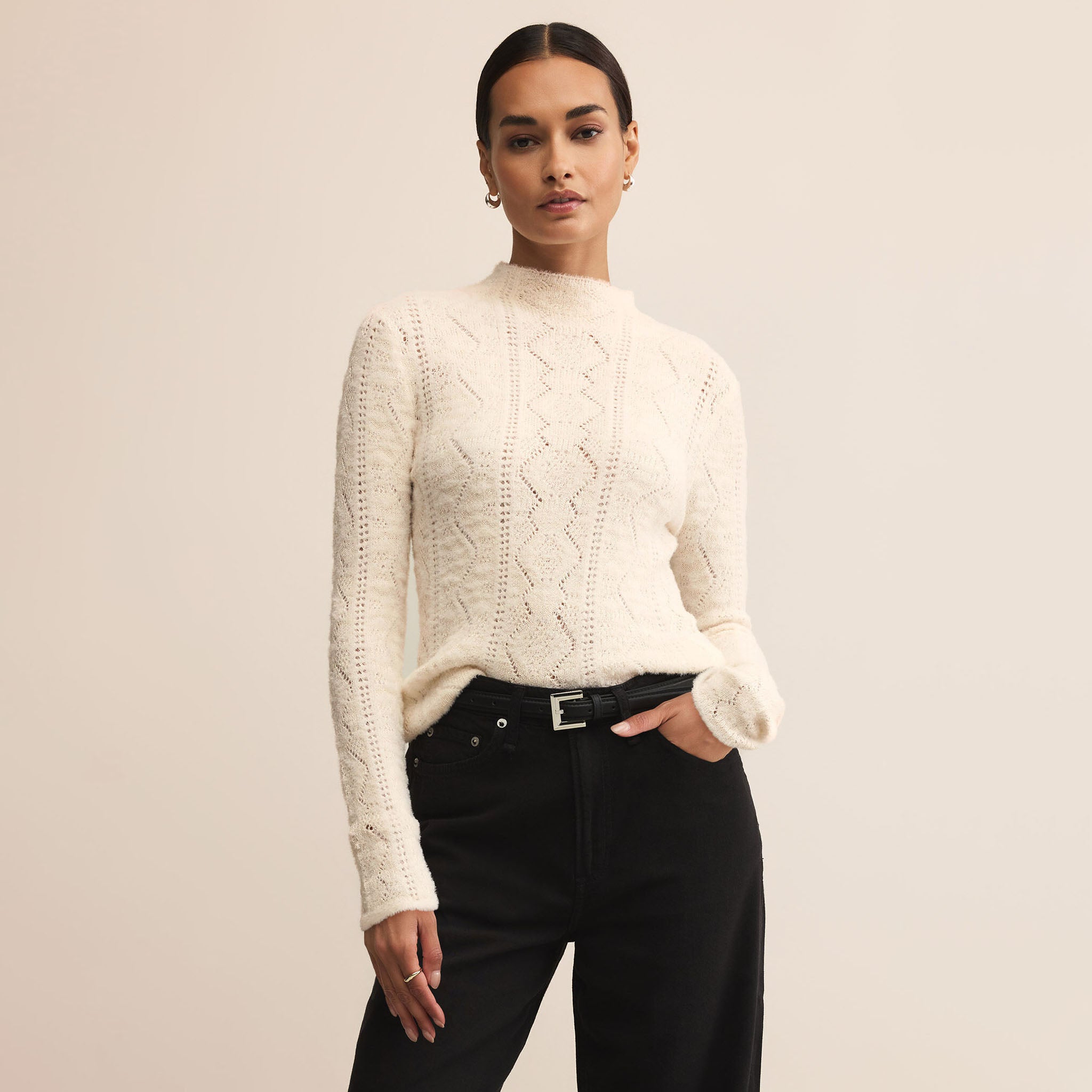 Woman wearing cream colored sweater with dark pants on a plain background.