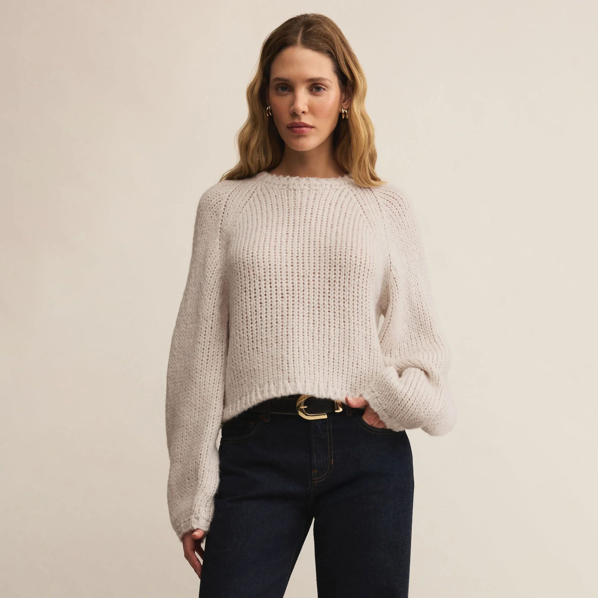 Woman wearing a beige sweater and dark jeans against a plain background