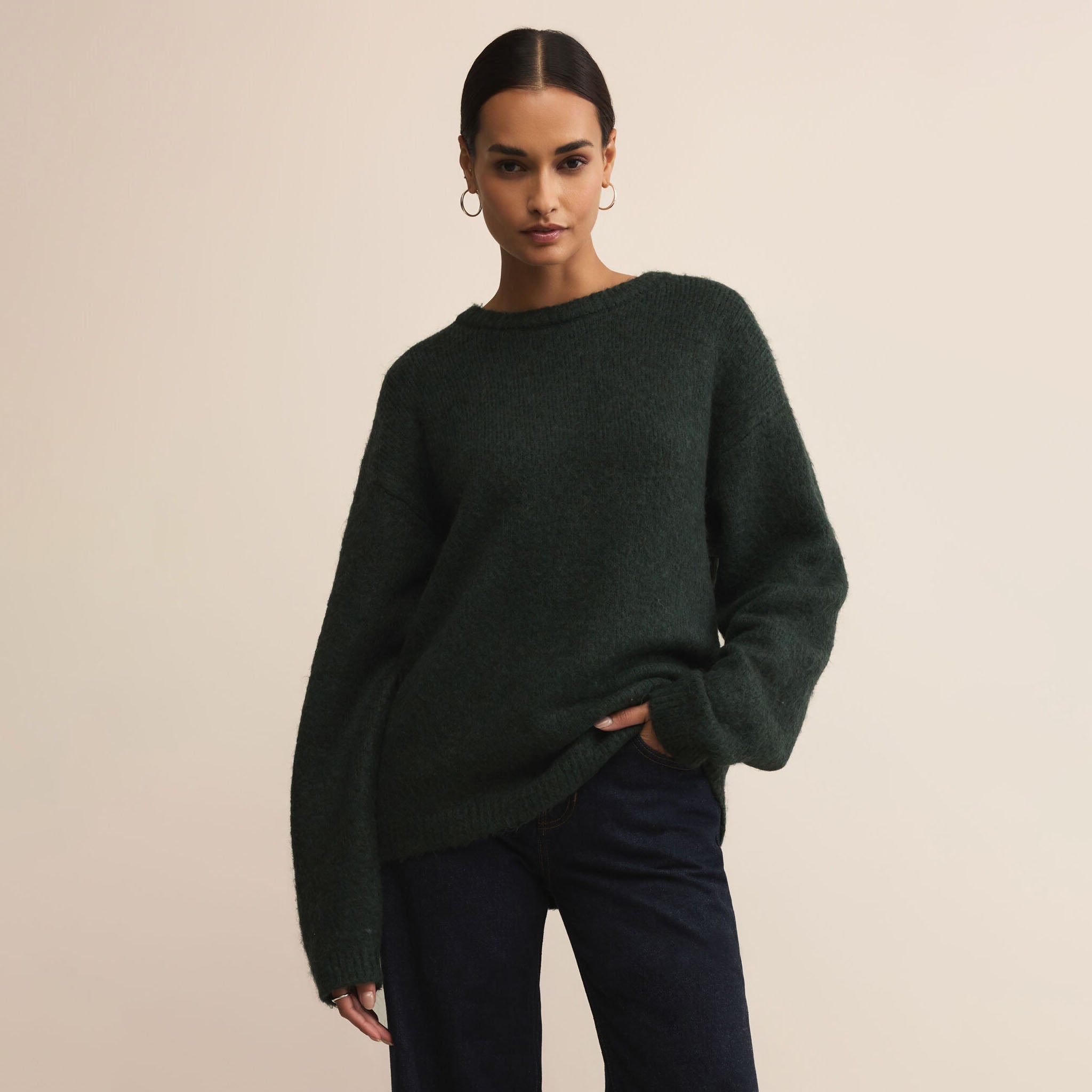 Woman wearing a green sweater and dark pants against a plain background.