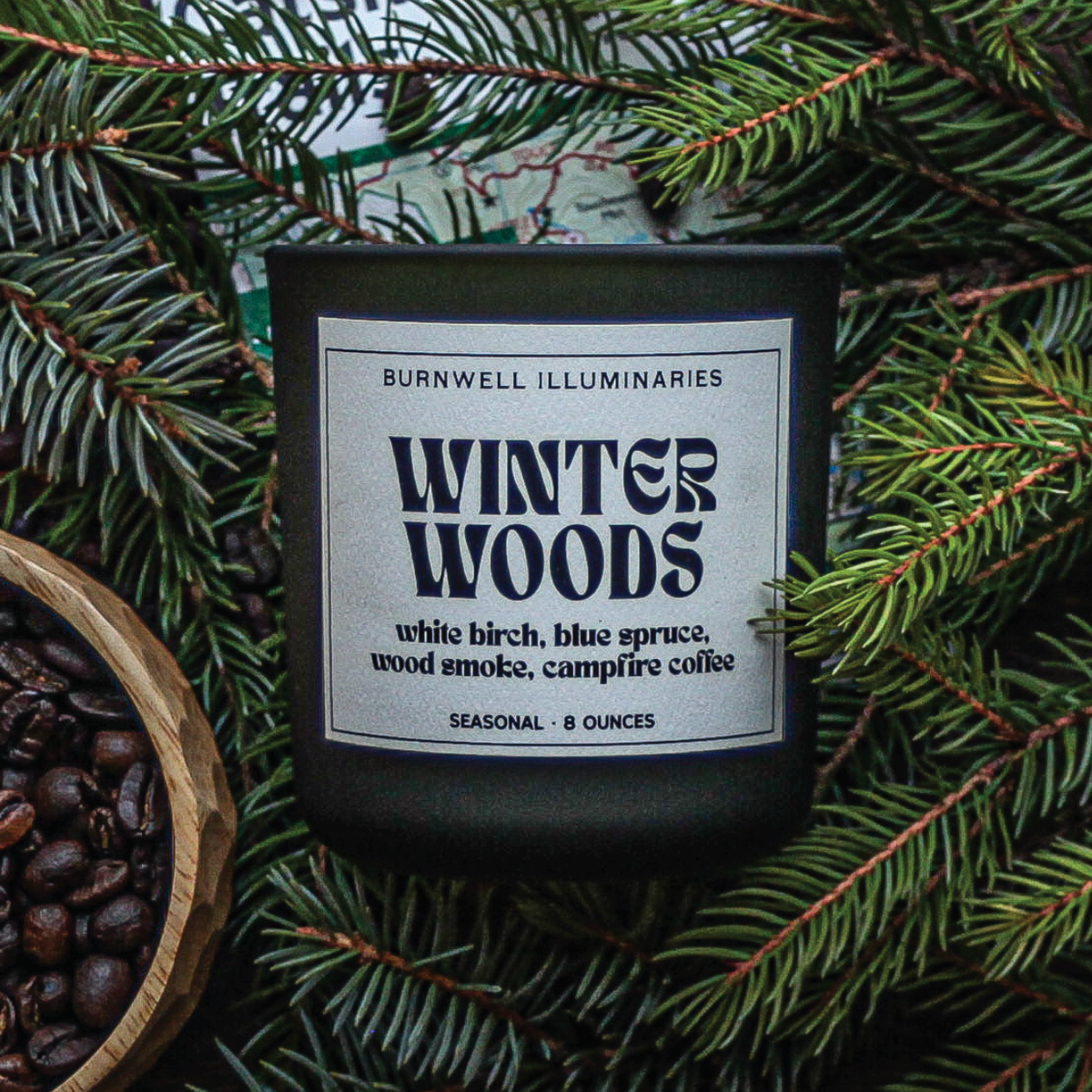 A dark glass candle jar labeled 'Winter Woods' by Burnwell Illuminaries, surrounded by pine branches and coffee beans, with a wooden bowl of coffee beans at the lower left.
