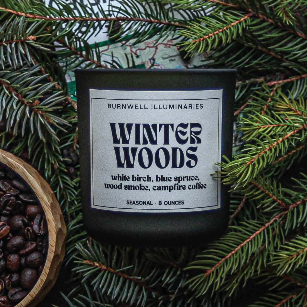 A dark glass candle jar labeled 'Winter Woods' by Burnwell Illuminaries, surrounded by pine branches and coffee beans, with a wooden bowl of coffee beans at the lower left.