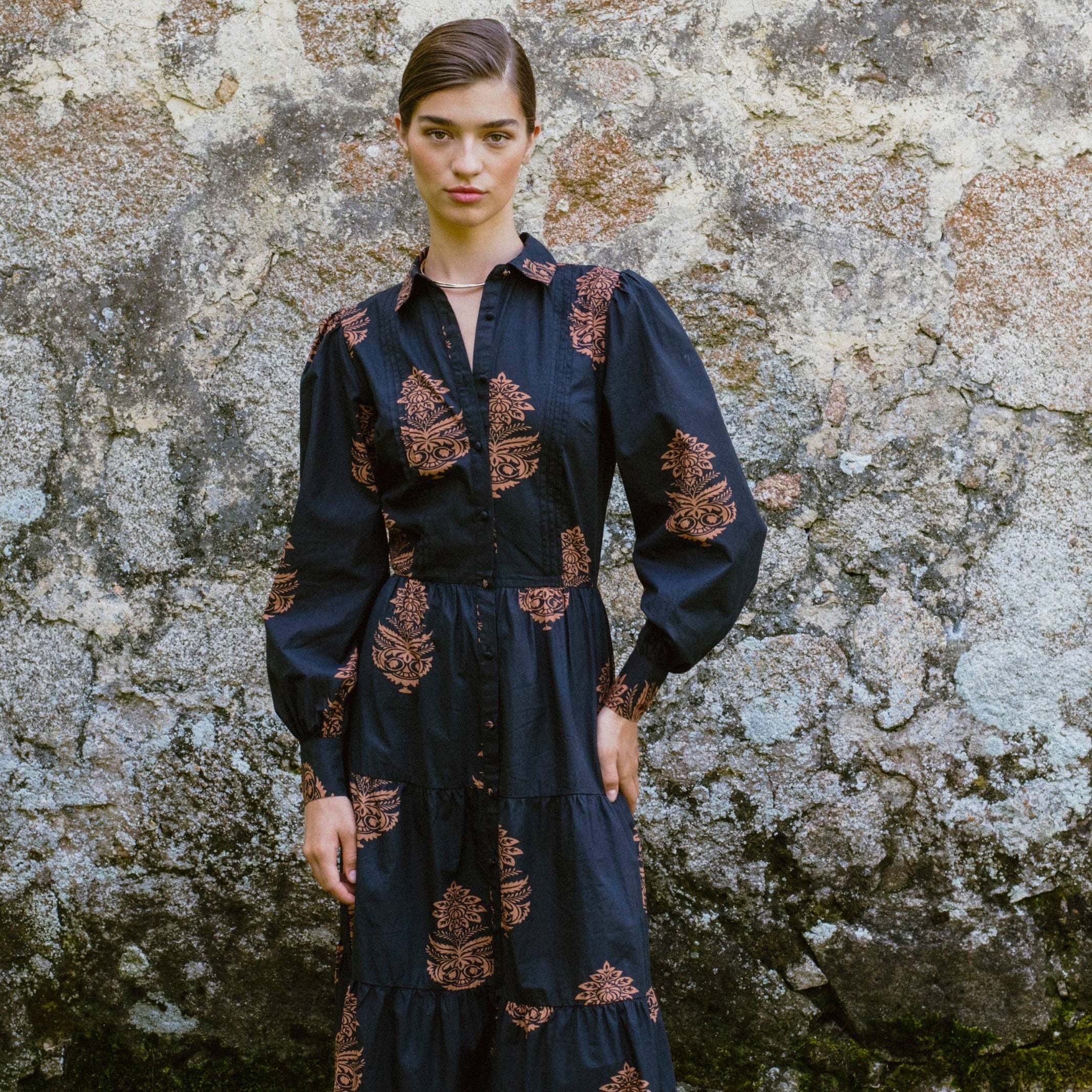 Woman wearing a dark blue dress with intricate patterns standing against a stone wall.