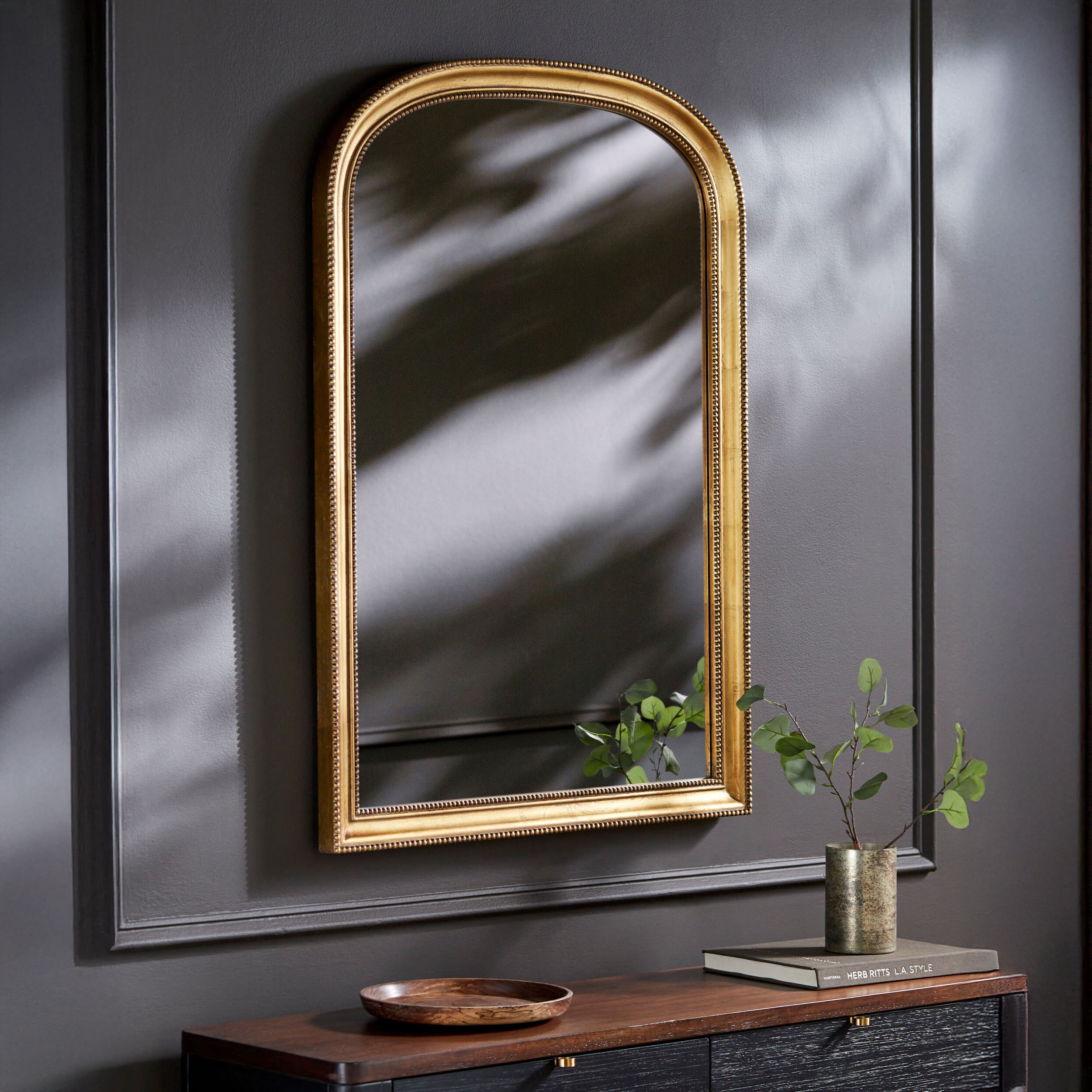 Nalanda Mantel Mirror in Antiqued Gold on a dark grey wall above a table at Addison West