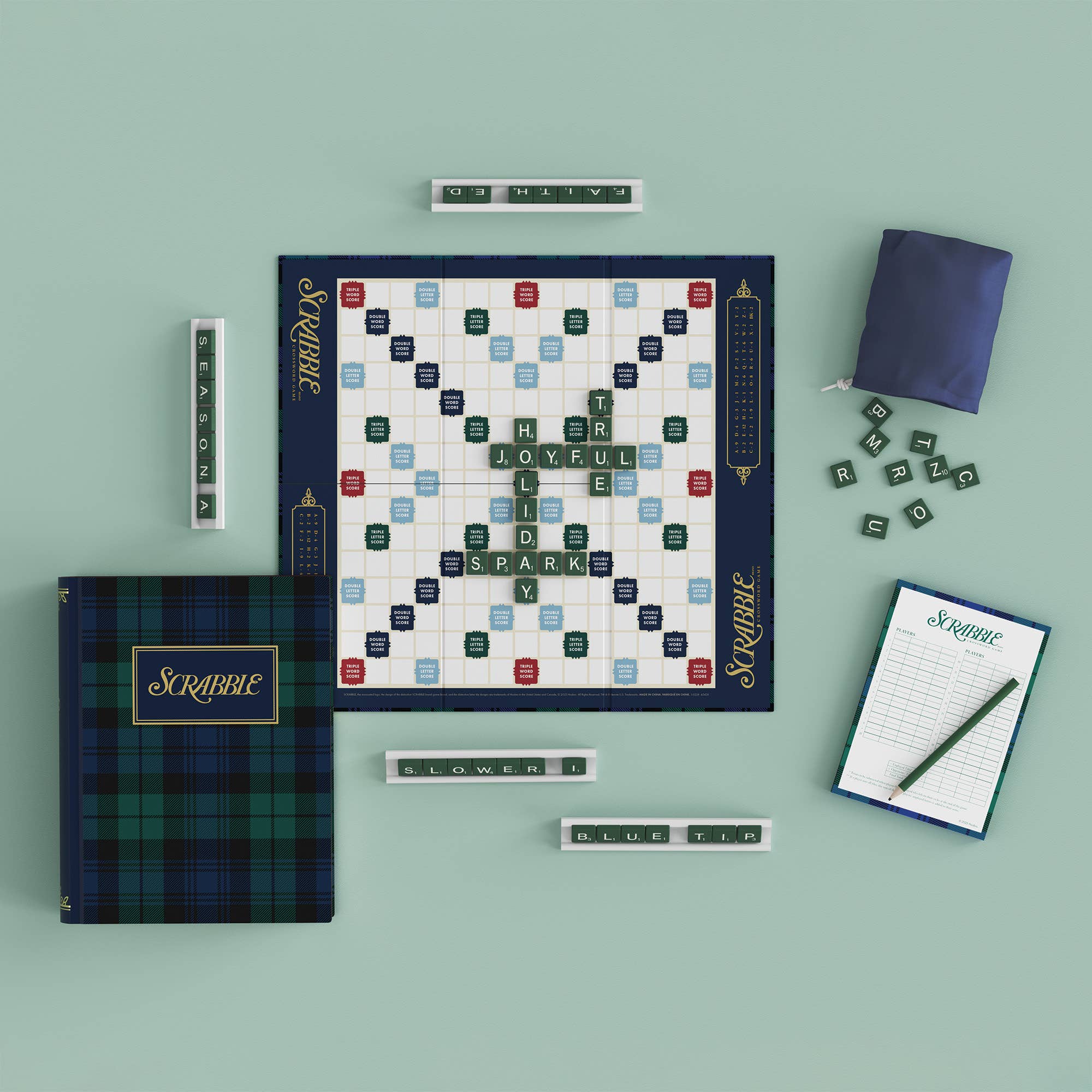 A flat-lay view of a Scrabble game set on a pale mint green background, including the game board, a plaid storage box, a blue bag, letter tiles, word tiles, a score pad, and a pencil.