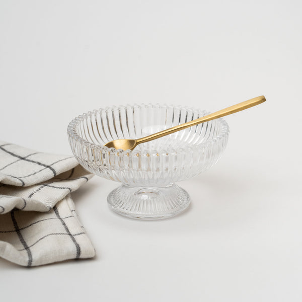 Pressed glass footed bowl with brass spoon and checked napkin on a white background