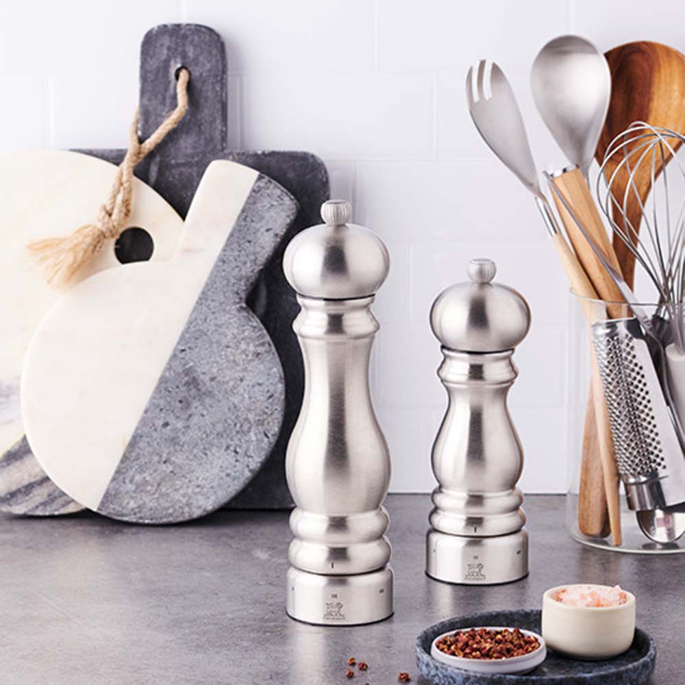 Set of two Peugeot Paris brand stainless mills on grey kitchen counter with various kitchen utensils