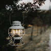 Barebones vintage white railroad lantern lit and hanging on a tree outside at dusk