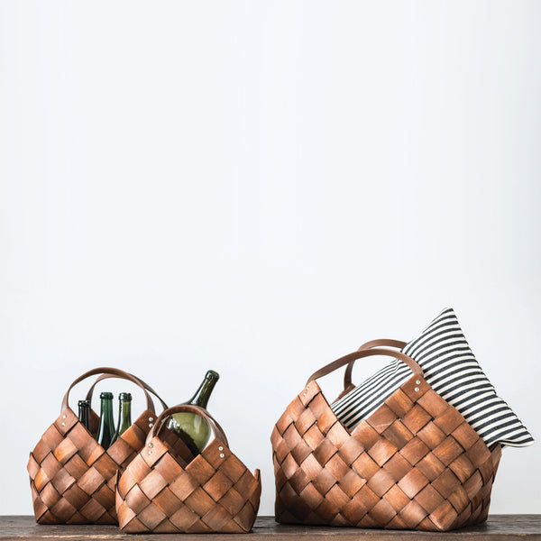 Three brown rectangular woven baskets with leather handles on a shelf with wine bottles