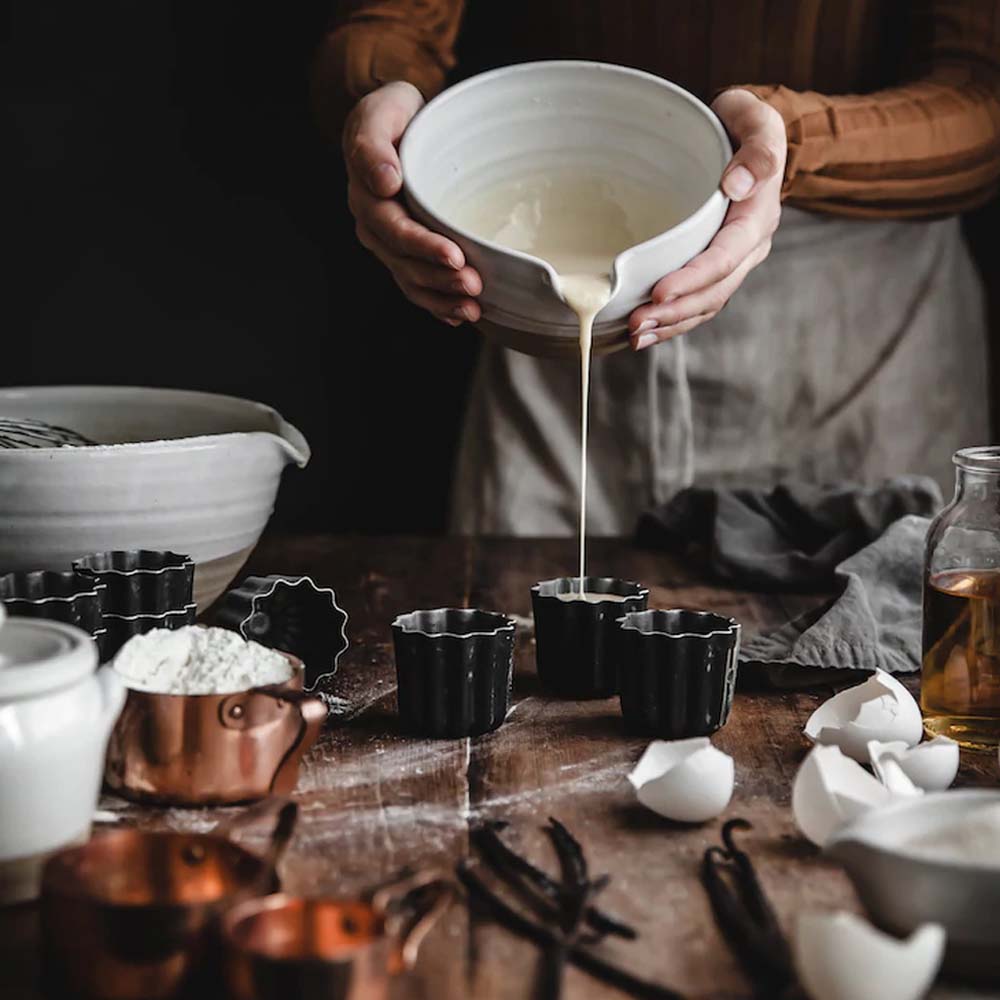 Woman pouring batter from a ceramic mixing bowl into baking molds on a table at Addison West