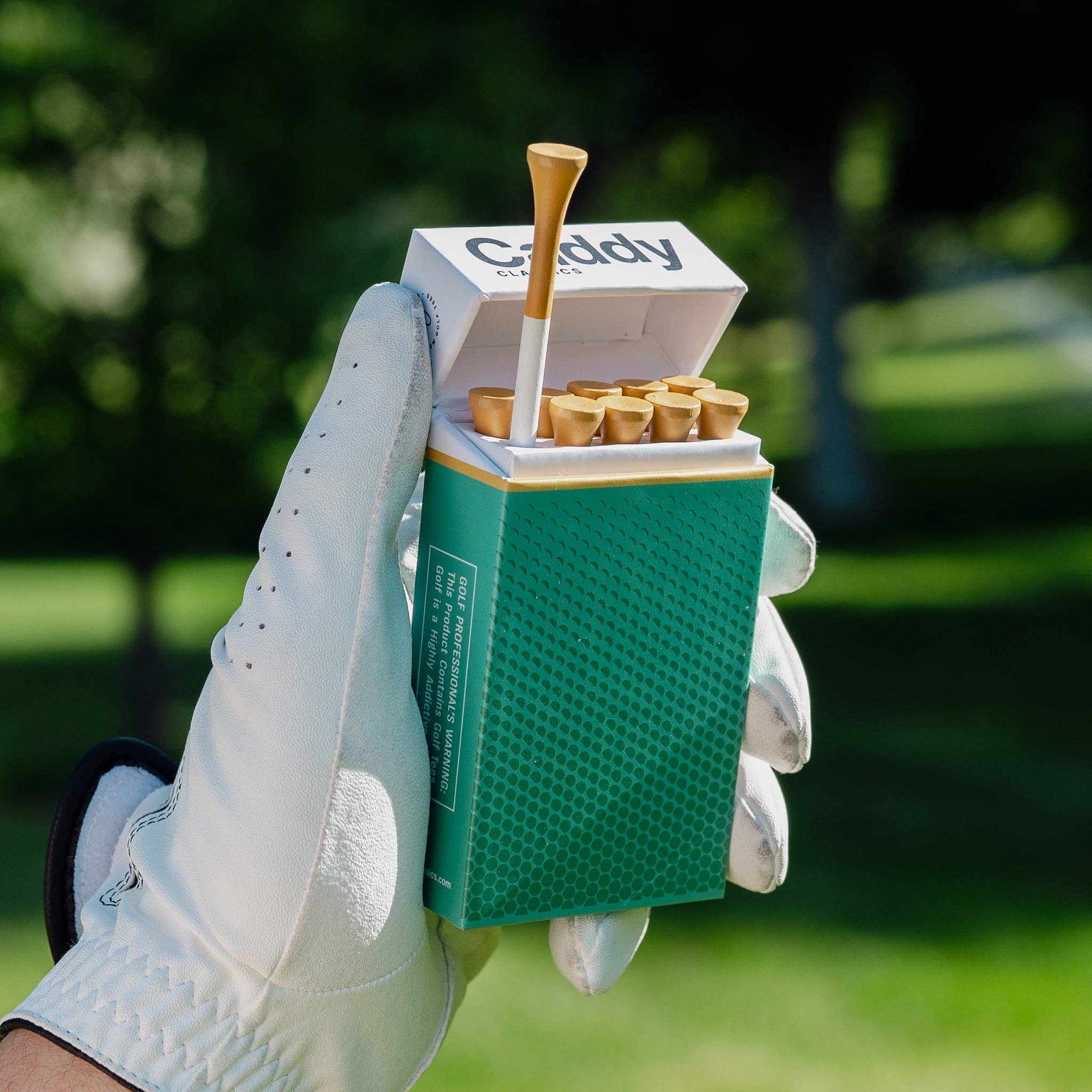 Green box of golf tees held by a gloved hand on a blurred golf course background