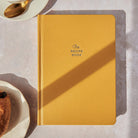 Yellow recipe book on a light surface with a spoon and dish in the background