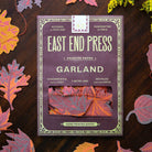 A package of East End Press Garland printed paper sheets lies on a dark wooden surface surrounded by colorful autumn leaves and small yellow flowers. The package is mauve-purple with cream text and border details, and a window near the bottom shows folded paper samples in pink, orange, and red tones.