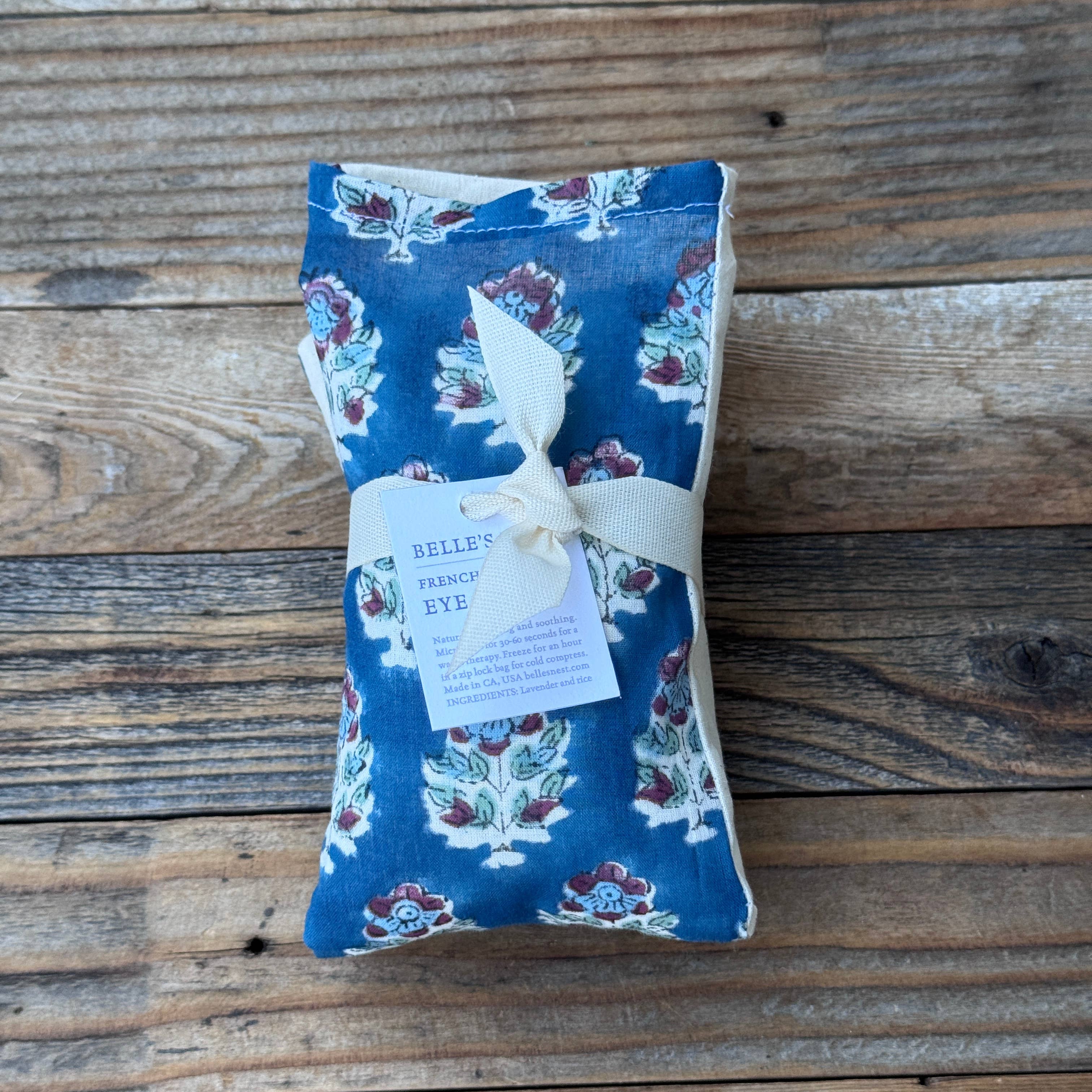 A blue floral eye pillow wrapped with a cream ribbon and a paper tag reading 'BELLE'S FRENCH YUKE', placed on a rustic wooden plank surface.