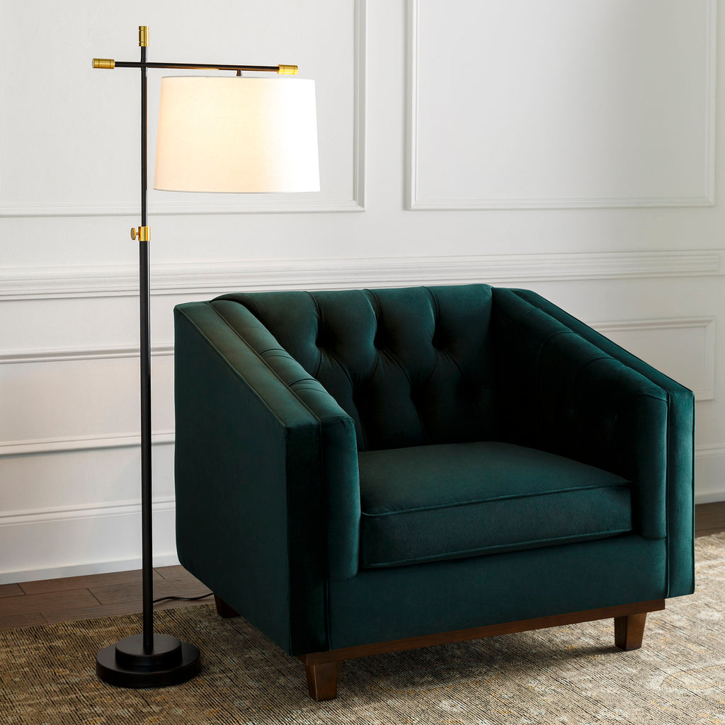 Surya Aberdeen floor lamp with black stand and stem and brass accents and a white linen shade next to a navy blue velvet chair in a living space with white walls