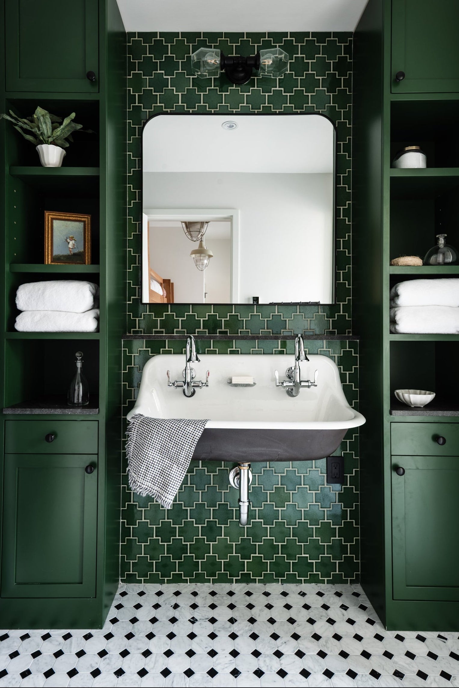 A styled bathroom interior featuring a white vintage-style pedestal sink with chrome faucets, set against a dark green geometric tiled wall and flanked by matching dark green built-in cabinets with shelves and folded white towels.
