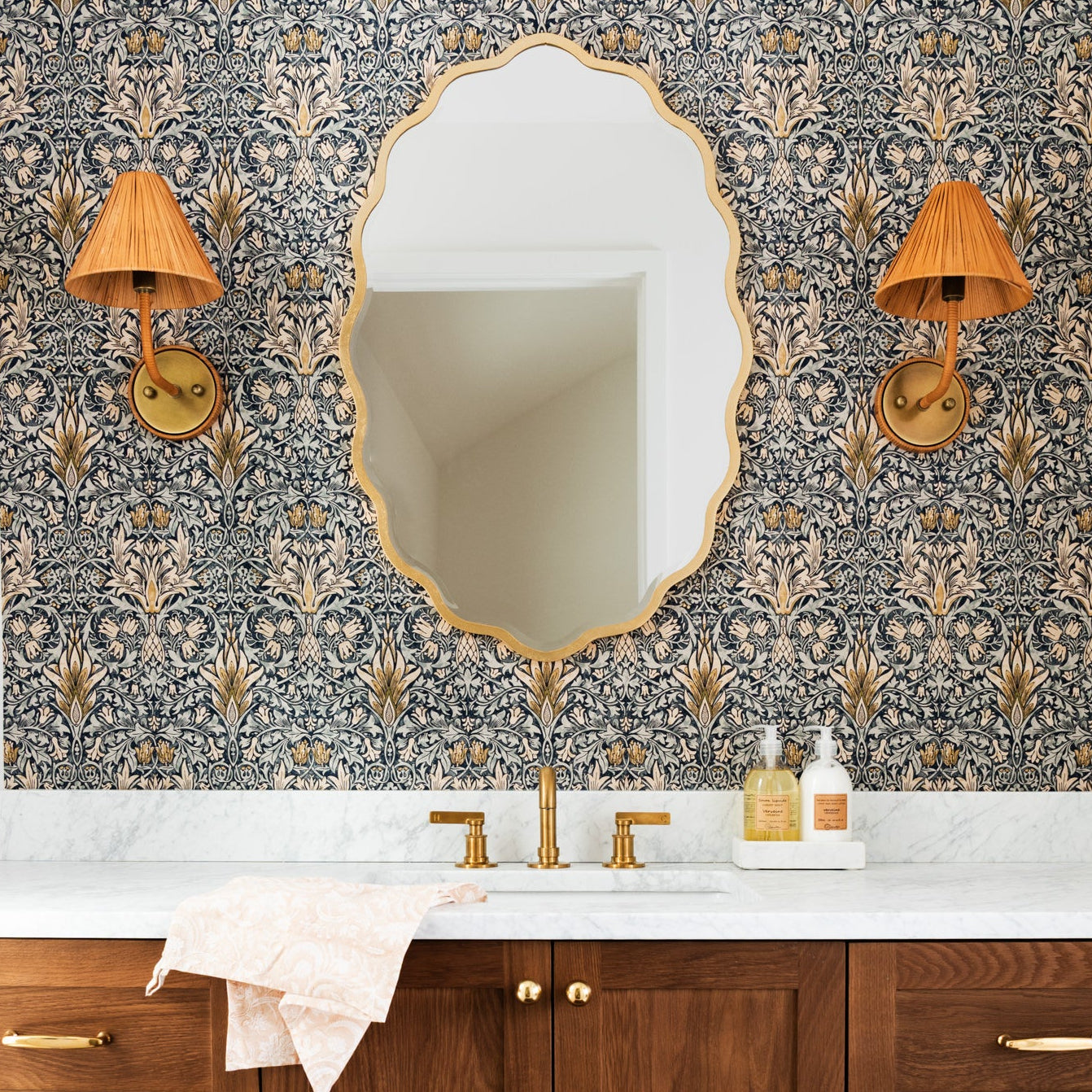 A styled bathroom vanity featuring a dark wood cabinet with brass handles, a white marble countertop, a large oval gold-framed mirror, and a decorative wallpapered wall with two gold sconces.