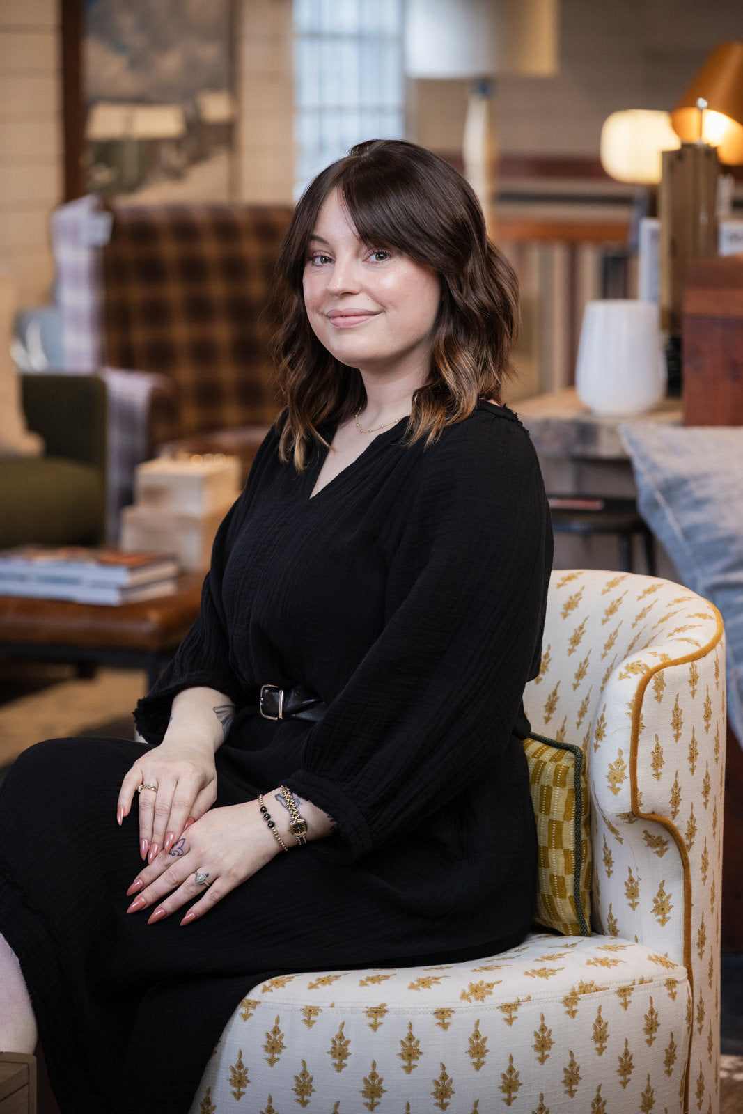 A woman with shoulder-length brown hair sits in a patterned armchair wearing a black long-sleeve dress with a thin belt. The setting is a warm, softly lit interior with home decor in the background.