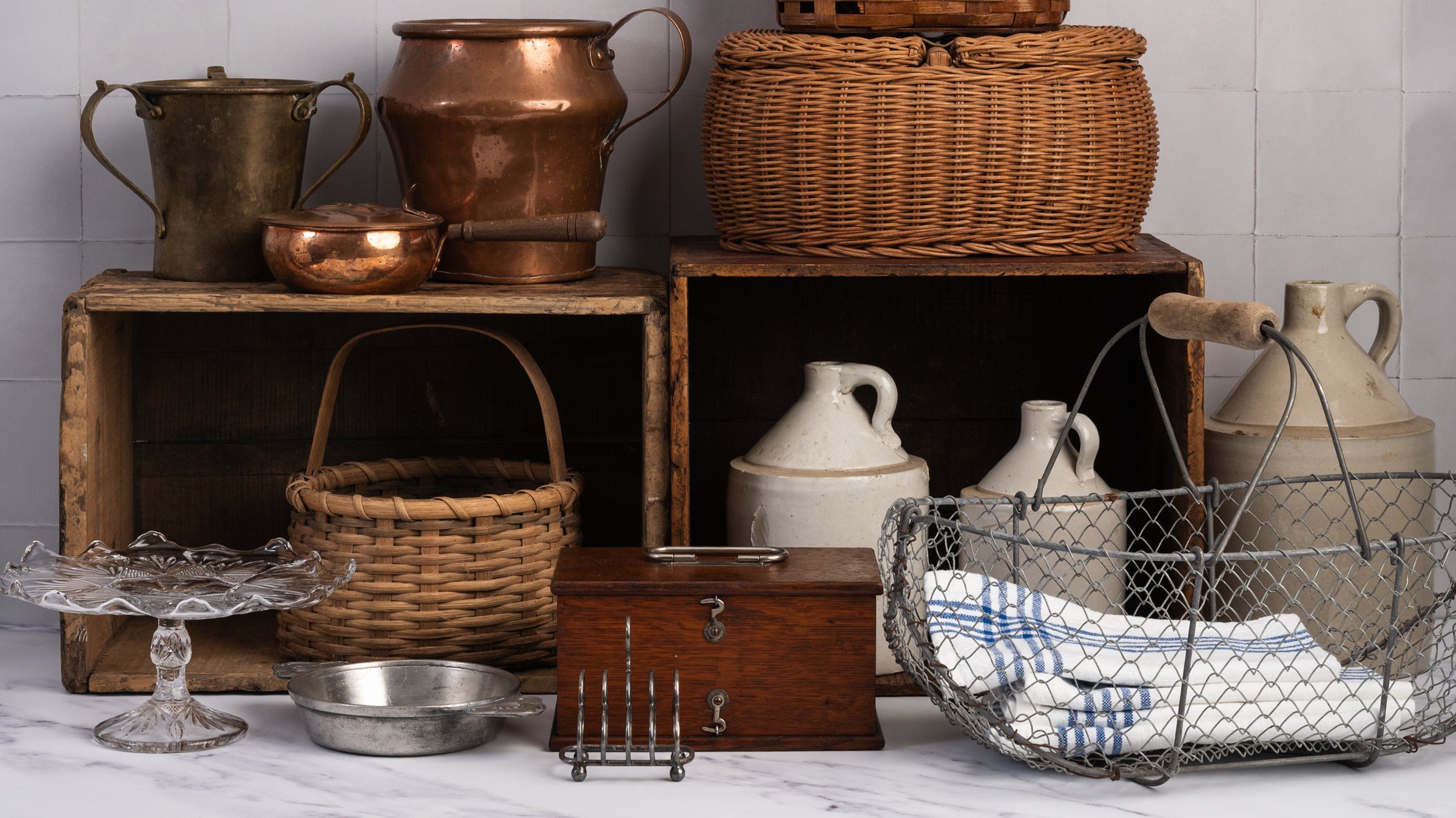 A styled collection of vintage rustic kitchen and storage vessels, including copper pots, woven wicker baskets, a metal wire basket with a blue-and-white striped cloth, ceramic jugs, and wooden crates on a marble countertop against a white tiled wall.