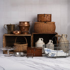 A styled collection of vintage rustic kitchen and storage vessels, including copper pots, woven wicker baskets, a metal wire basket with a blue-and-white striped cloth, ceramic jugs, and wooden crates on a marble countertop against a white tiled wall.