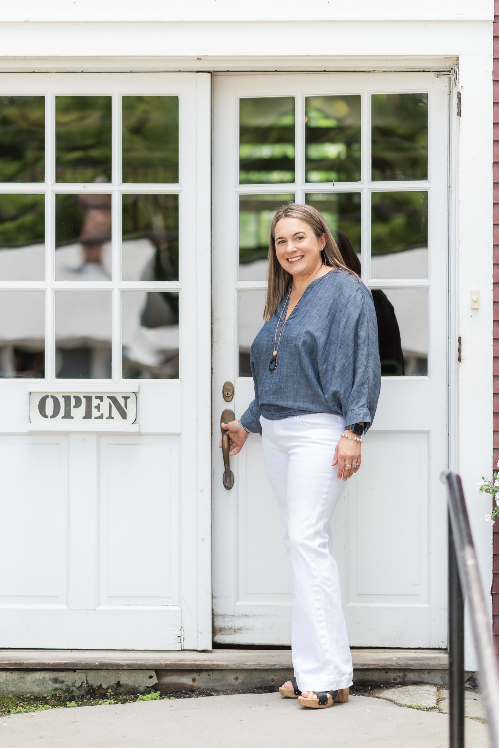 A woman stands in front of a white storefront door with glass panes, wearing a blue long-sleeve blouse and white pants. The door has an 'OPEN' sign and a metal handle.