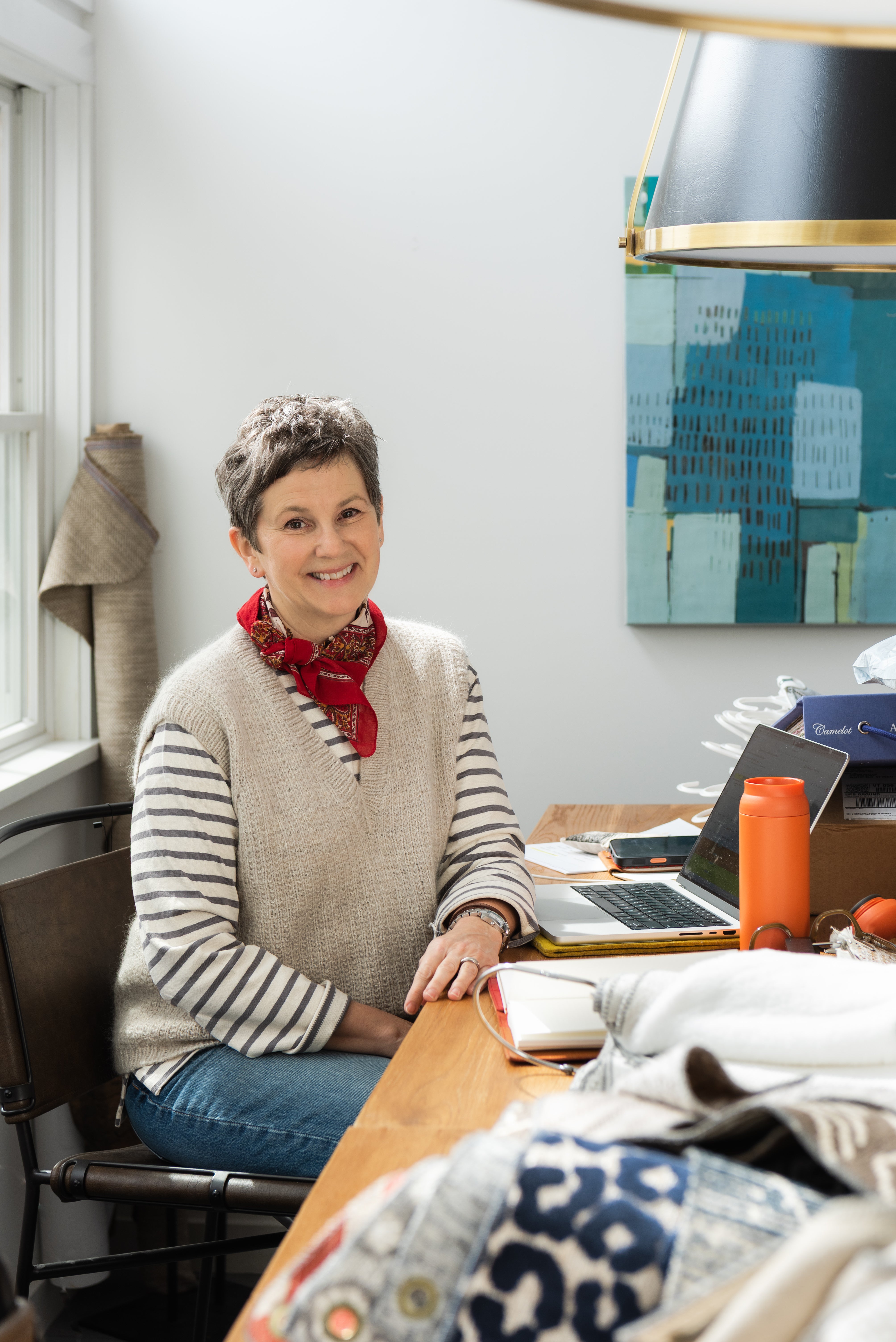 A smiling woman with short gray hair sits at a wooden desk in a bright room, wearing a beige knit sweater vest over a black-and-white striped long-sleeve shirt, blue jeans, and a red patterned neck scarf. A laptop, orange bottle, and patterned fabric are visible on the desk.